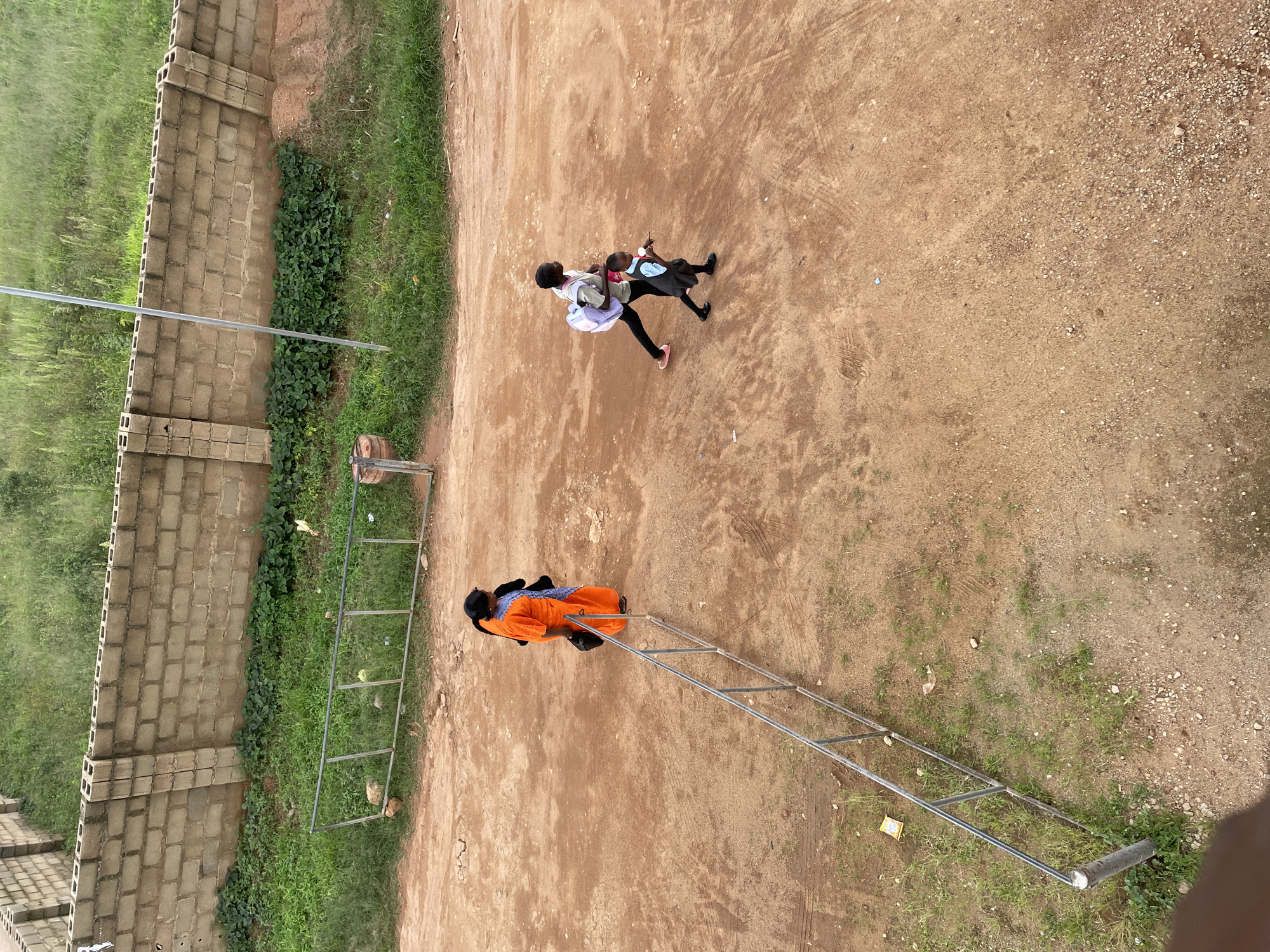 An overhead shot shows a dirt path with a brick wall and tall grass in the background, likely in the city of Jos, Nigeria. To the left, a woman in an orange garment stands, holding a black bag. To the right, two children walk together, the taller one carrying a light purple backpack, and the shorter one, in a school uniform, holding a small white object. A metal railing runs diagonally across the scene.