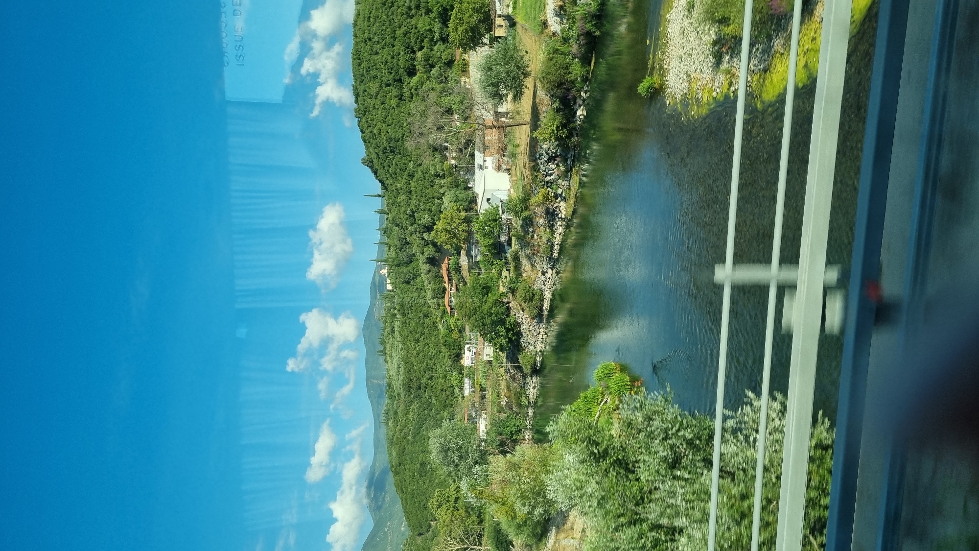 The image shows a wide, calm river flowing through a green, hilly landscape. On the far bank of the river, there are several buildings, some with red-tiled roofs, nestled amongst trees and vegetation. Further up the hill, more scattered buildings and cypress trees are visible. The sky is a bright blue with scattered white clouds. In the foreground, a white metal railing of a bridge or overpass runs horizontally across the lower portion of the image. The river reflects the sky and surrounding greenery. The scene appears to be located in a rural or semi-rural area, consistent with the topography of Δημοτική Ενότητα Ευρυμενών, Greece. No people or animals are clearly visible.