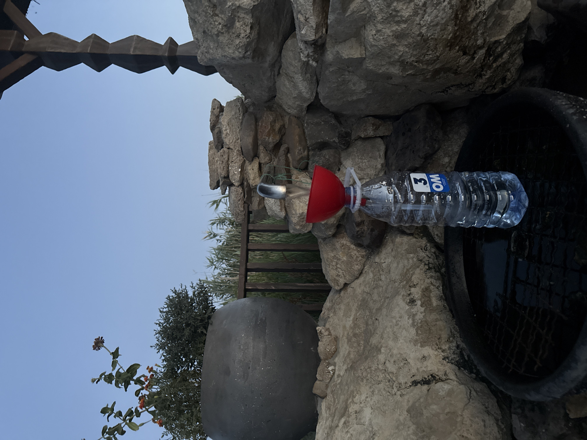 The image depicts an outdoor scene with a water bottling setup. A clear plastic water bottle, partially filled with water, stands on a dark, circular surface, possibly a grate within a basin. A red funnel is attached to the top of the bottle, and a thin metallic spigot extends into the funnel, from which water is flowing. The background consists of a stone wall and natural elements, including rocks and some greenery. The sky above is a clear, light blue, suggesting it is either early morning or late evening. The environment appears to be somewhat rustic. There are no people visible in the image. The scene does not appear to be located in Cimișlia, Moldova, based on common visual cues associated with that region.