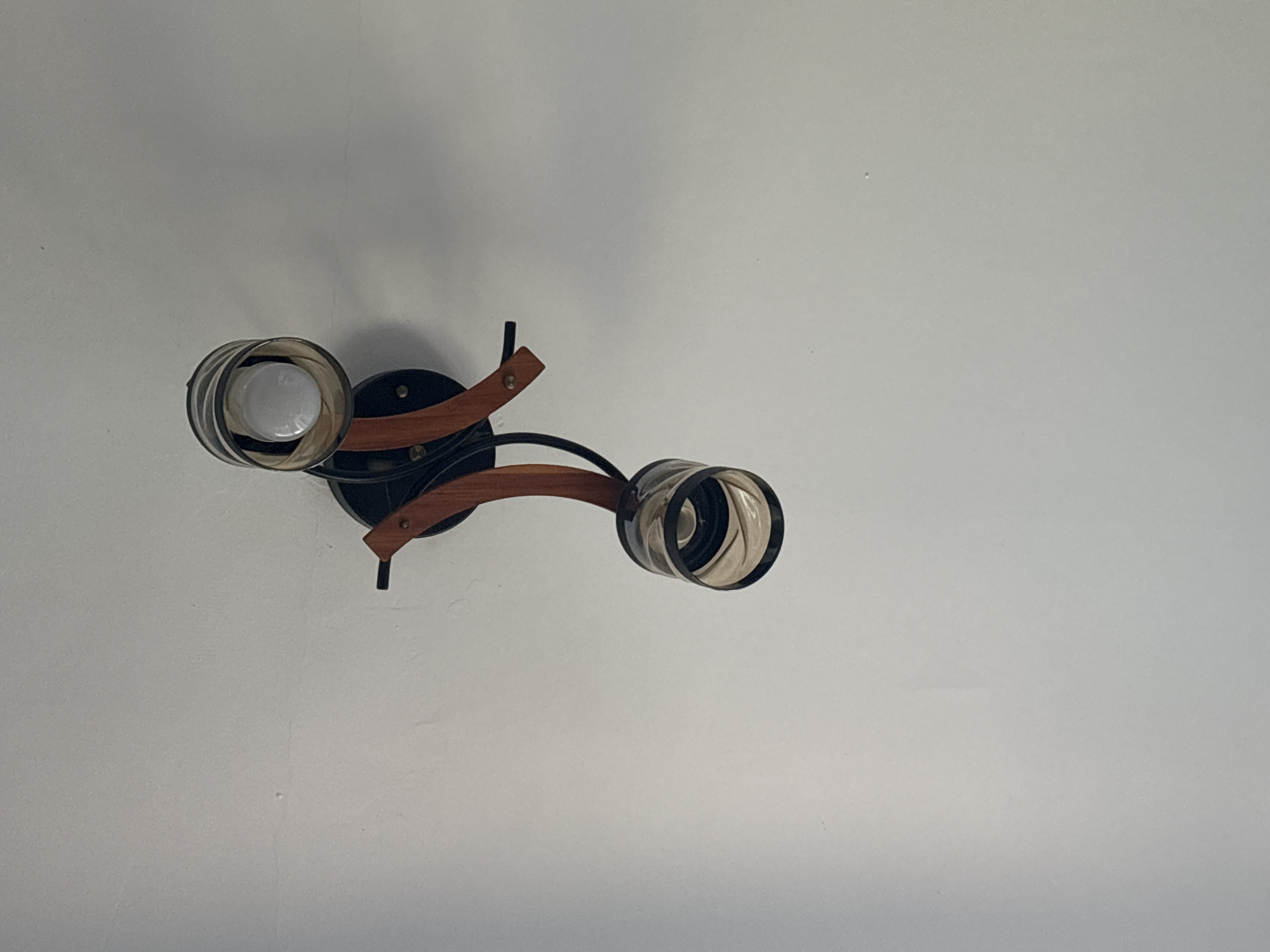 The image shows a close-up, overhead view of a semi-flush mount ceiling light fixture against a white ceiling. The fixture has a black base and two arms that curve downwards and outwards. Each arm supports a cylindrical glass shade with a ribbed interior. One shade contains a white LED bulb, which is illuminated. The other shade is empty. The fixture is designed in a modern style with accents of what appears to be natural wood. The ceiling is plain and shows some minor imperfections, such as small dots and faint lines. The lighting in the image suggests it was taken indoors during daylight hours, likely from within a room. There are no people or other objects visible in the frame.