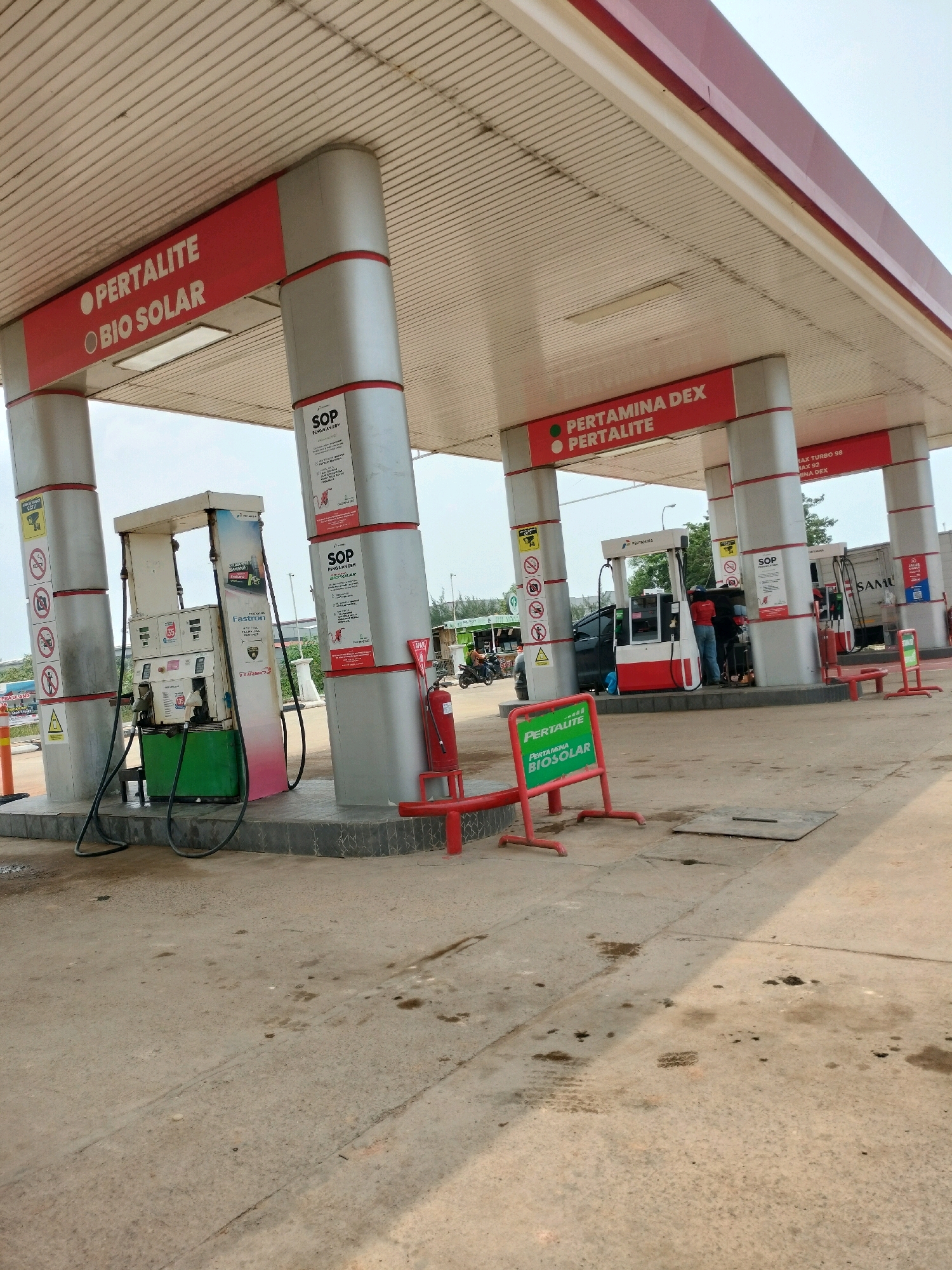The image depicts an outdoor view of a Pertamina gas station in Batam, Indonesia, on an overcast day. The gas station features a red and white canopy supported by gray pillars. Several fuel pumps are visible, some with visible branding such as "Pertalite" and "Bio Solar." One pump has a green base with pink accents and black hoses. Another pump has a red and white design. A black car is parked at one of the pumps, with an attendant in a red shirt and blue jeans attending to it. A person on a motorcycle is visible in the background. Red safety barriers are placed in front of the pumps. Green and red signs with white text indicating fuel types are present. The ground is paved with concrete, showing signs of wear and some oil stains. The overall environment appears to be a typical urban gas station setting.