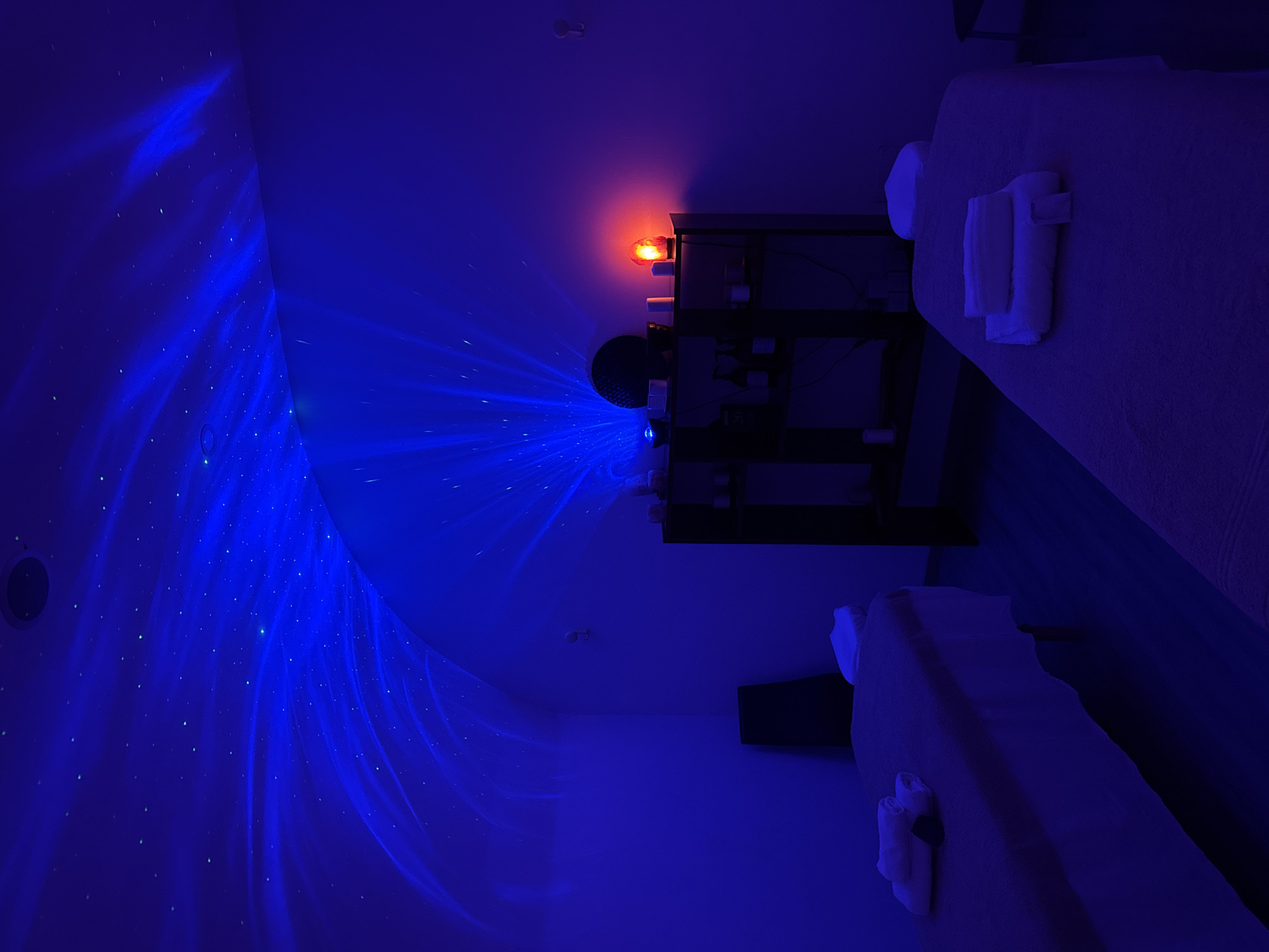 The image depicts a dimly lit room with two massage tables, each covered in a light-colored sheet and topped with folded white towels. The room is illuminated by a starry sky projector, casting a vibrant blue light and numerous small white dots across the walls, creating a celestial effect. A red-orange light emanates from a fixture above a dark shelving unit, which is also visible in the scene, adorned with various items, possibly spa products. The overall ambiance is serene and relaxing, suggesting a spa or therapy setting. The time of day is likely evening or night due to the dim lighting and the focus on artificial light effects. The location is specified as Isola delle Femmine, Italy. No people are visible in the image, and there is no visible text.