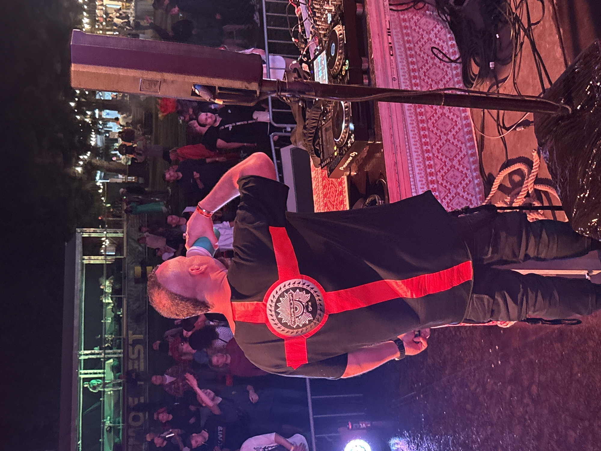 An outdoor music festival is in progress at night in Polevaya, Russia, with a DJ performing on a raised stage. The DJ, a man with a mohawk hairstyle and a black t-shirt with a red cross emblem, is seen from behind, drinking from a teal cup. He is wearing dark cargo pants. The DJ booth features Pioneer DJ equipment and is adorned with a decorative red and white patterned cloth. In the background, a crowd of people are gathered, illuminated by stage lighting and string lights in the trees. A large speaker is positioned next to the DJ. The ground appears wet, possibly from rain or spilled drinks. A metal fence separates the stage area from the audience.
