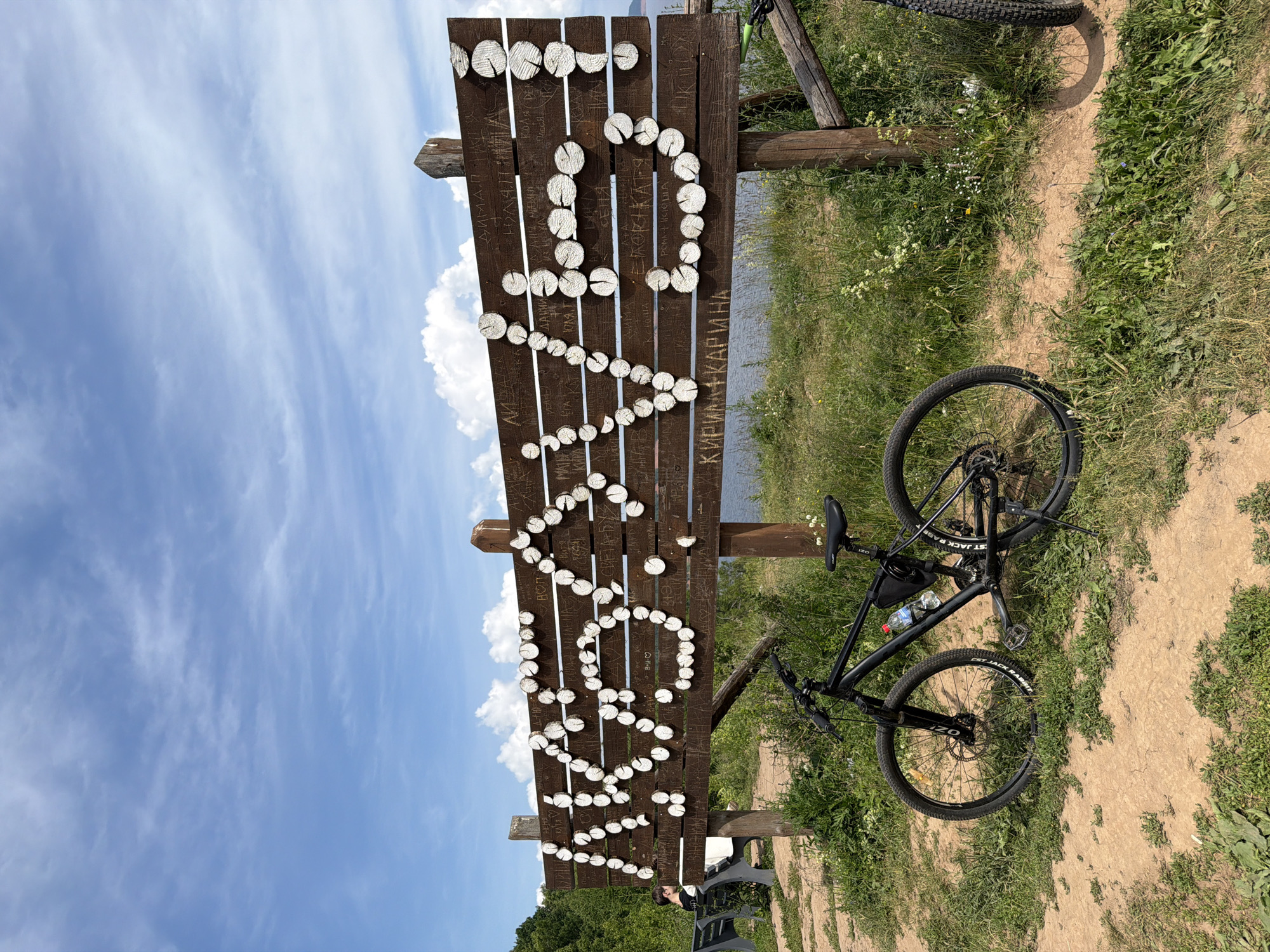 The image shows a wooden sign with the word "КОСТОВАТЫ" written on it in Russian, using slices of wood as letters. The sign is brown and weathered, with some graffiti scratched into its surface. To the left of the sign, a person is sitting on a park bench. In front of the sign, a black bicycle is parked on a dirt path, with a water bottle and a small bag attached to its frame. The background shows lush green grass, trees, and a body of water, suggesting an outdoor location. The sky is blue with scattered white clouds.