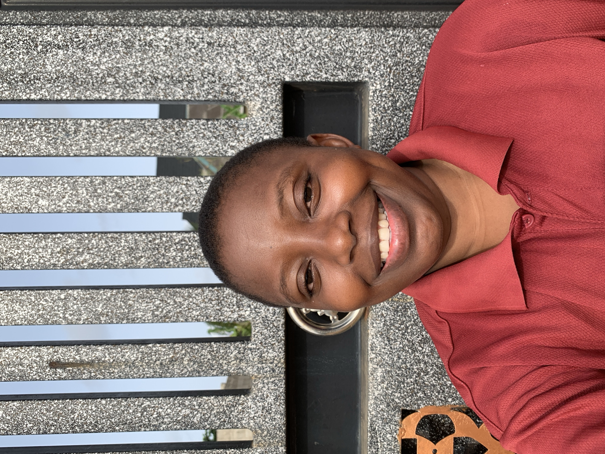 A medium shot of a dark-skinned person smiling with a reddish brown collared shirt. The person is positioned in front of a gray gate with horizontal mirrored bars and black vertical bars, along with black and gold accents at the bottom. The background includes what looks to be some greenery and hints of buildings. The location is speculated to be Ikorodu, Nigeria.