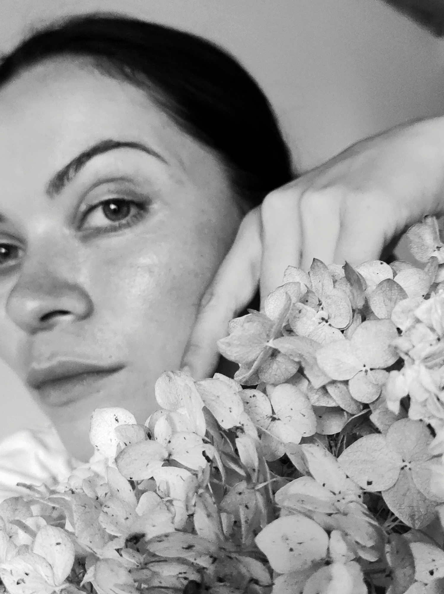 This is a black and white, close-up, portrait-style photograph of a woman. The woman is positioned on the left side of the frame, with her face partially obscured by a cluster of dried flowers in the foreground. Her left eye, eyebrow, nose, and lips are visible. Her left hand rests on her cheek, near the flowers. The background is a plain, light-colored surface. The lighting appears soft, creating a gentle mood. The image quality suggests it may be a photograph from the early 2000s or later, given the digital artifacting. The setting appears to be indoors, with a neutral, unadorned backdrop. The woman's expression is subtle, possibly contemplative or serene. No text is visible in the image. The location context provided is "Foki, Russia," which is not directly discernible from the visual content of the image itself.