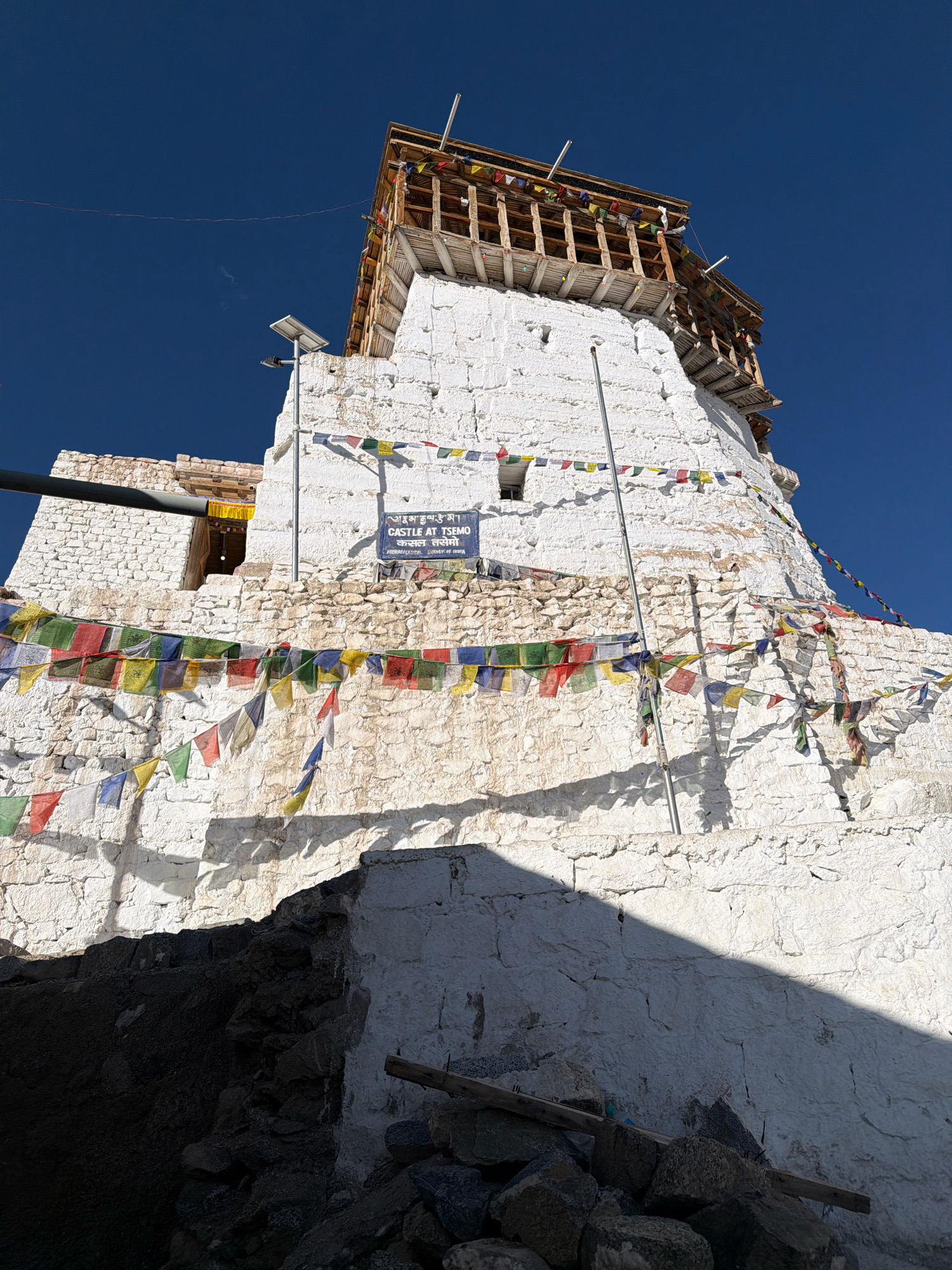 This is a low-angle, wide shot of the Tsemo Castle in Leh, India, under a clear, deep blue sky. The white-washed stone structure of the castle dominates the frame. Prayer flags in various colors, including red, blue, yellow, green, and white, are strung across the façade of the castle, fluttering in an unseen breeze. The building features wooden balconies and tiered roofs, with some parts appearing weathered and aged. A sign is affixed to the wall, with text in both Tibetan script and English, reading "CASTLE AT TSOYO". The lower portion of the image shows a rough pile of dark rocks and debris, casting long, distinct shadows across the white walls. A solar-powered street light is mounted on a pole against the castle wall.