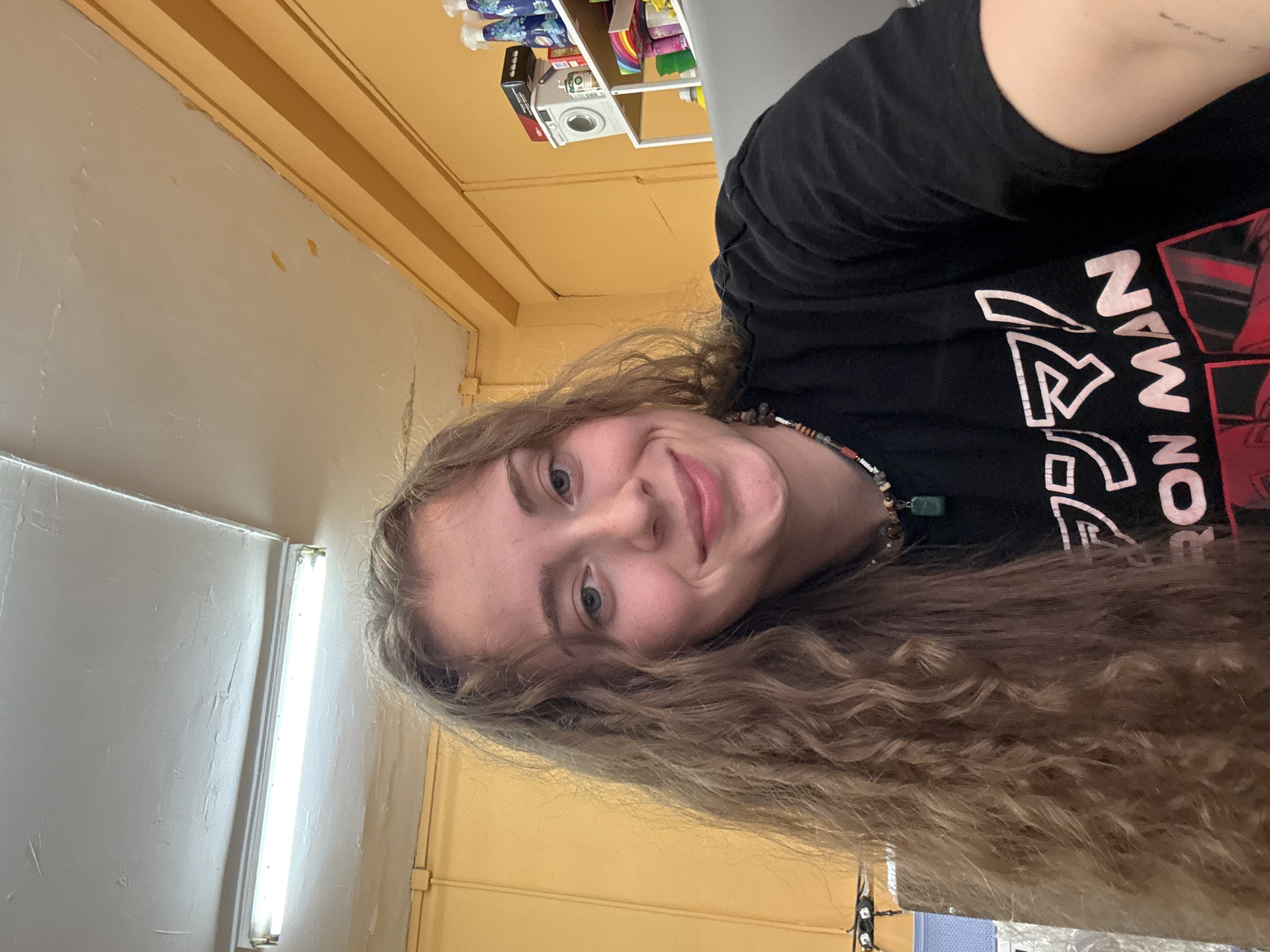 This is a portrait-style selfie of a young woman with long, curly, light brown hair. She is wearing a black t-shirt with white and red graphics. A beaded necklace with a green pendant is visible around her neck. Her facial expression is a friendly smile.
The background suggests an indoor setting, possibly a room with yellow walls and white fluorescent lighting. There are shelves in the upper background with various items, including what appears to be cleaning supplies and a small white appliance. The overall impression is casual and informal.
The image appears to have been taken during the daytime, based on the lighting. No specific weather conditions are discernible. The location context of Sofia, Bulgaria, does not provide specific visual cues in this particular image, but it places the subject geographically. There is no discernable text in the image.