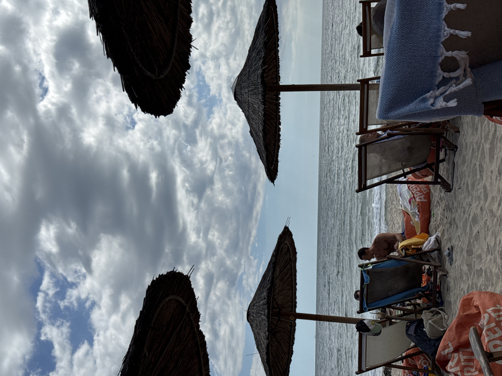 A wide shot captures a beach scene on a cloudy day. In the foreground, beach chairs and large beanbag-style cushions are arranged on the sand. Two large thatched umbrellas provide shade. In the midground, a calm sea stretches to the horizon. A man is seated on a chair, looking out at the sea, with another person visible behind him. A blue towel is draped over a chair in the right foreground. The scene appears to be at a beach resort, possibly in the Varna, Bulgaria area, given the general ambiance and the common presence of such amenities in coastal Bulgarian cities.