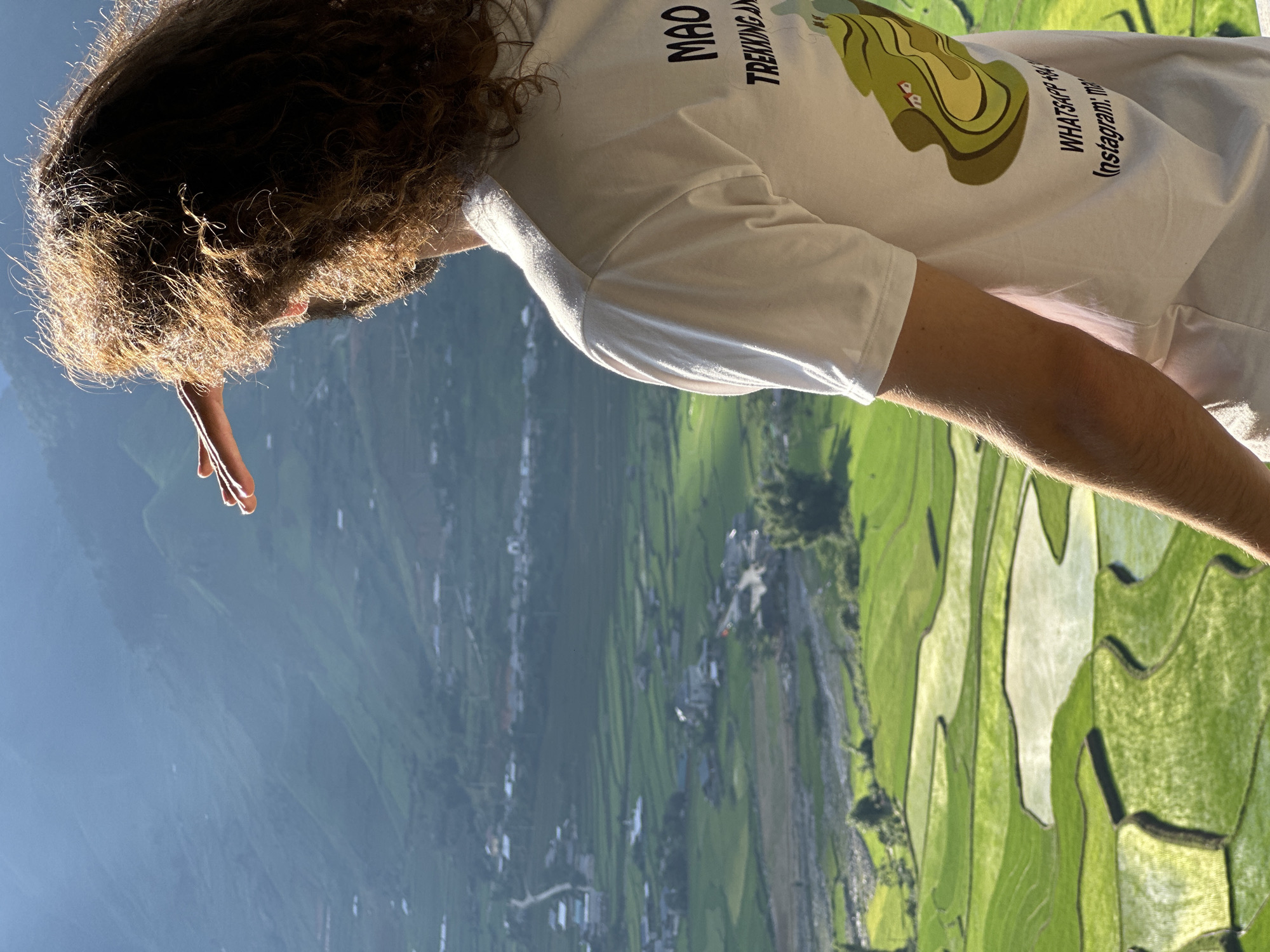 A person with long, curly, brown hair is viewed from behind, standing outdoors and looking into a vast valley in Tú Lệ, Vietnam. The individual, whose face is not visible, is wearing a white short-sleeved t-shirt. Their right hand is raised to shield their eyes from the sun. The t-shirt features text on the back: "MAO TREKKING AND" on the upper right side, and "WHATSAPP +84" and "Instagram: ma" (partially visible) towards the bottom right. An illustrated graphic depicting green terraced fields with small houses is also printed on the back.
The background reveals a expansive landscape characterized by lush green terraced rice paddies, some filled with reflective water, cascading down hillsides. In the midground, a small village with scattered houses and a winding road is visible along a river or stream. Distant mountains rise, creating a hazy, layered appearance under what appears to be bright sunlight from the left, highlighting the person's hair and shoulder.