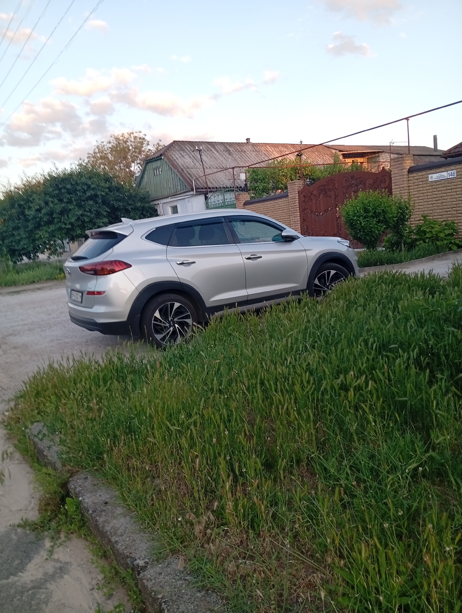 The image shows a silver Hyundai Tucson parked on a dirt road next to a curb with overgrown grass. In the background, there are houses with tiled roofs, a brick fence, and a large metal gate. The sky is partly cloudy. There are no people visible in the image. The scene appears to be in a residential area.