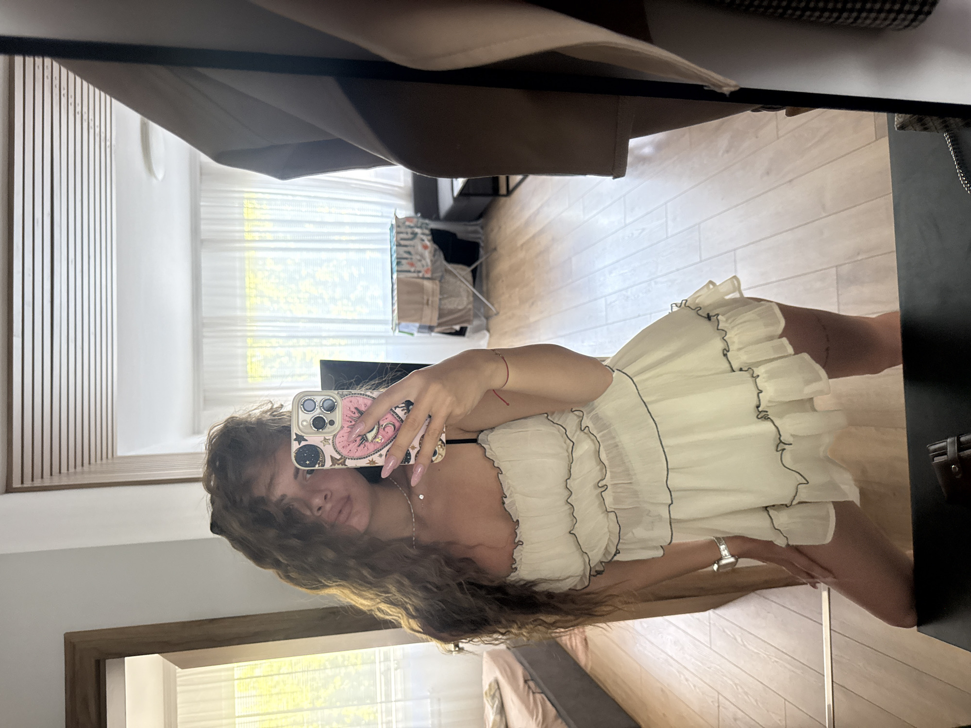A woman stands in front of a mirror in an indoor location, likely a residence, in the city of Unknown, Unknown. The woman is holding a phone, using it to capture her image in the mirror. She is wearing a white dress with ruffled edges. Her long brown hair falls over her shoulder and she wears a necklace and a watch. The background shows a room with light wood flooring, a window with sheer curtains, and furniture partially visible. There's a clothes drying rack in the distance.