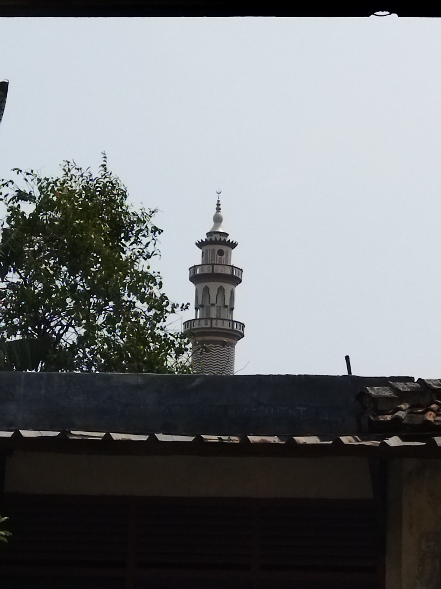 A minaret tower of a mosque rises above a dark roofline, partially obscured by a large green tree on the left. The minaret has multiple levels, with arched windows and decorative trim. The sky is overcast. The scene appears to be in a residential or mixed-use area, as indicated by the roof tiles and the suggestion of buildings in the foreground. This image was taken in Paku Haji, Indonesia.
