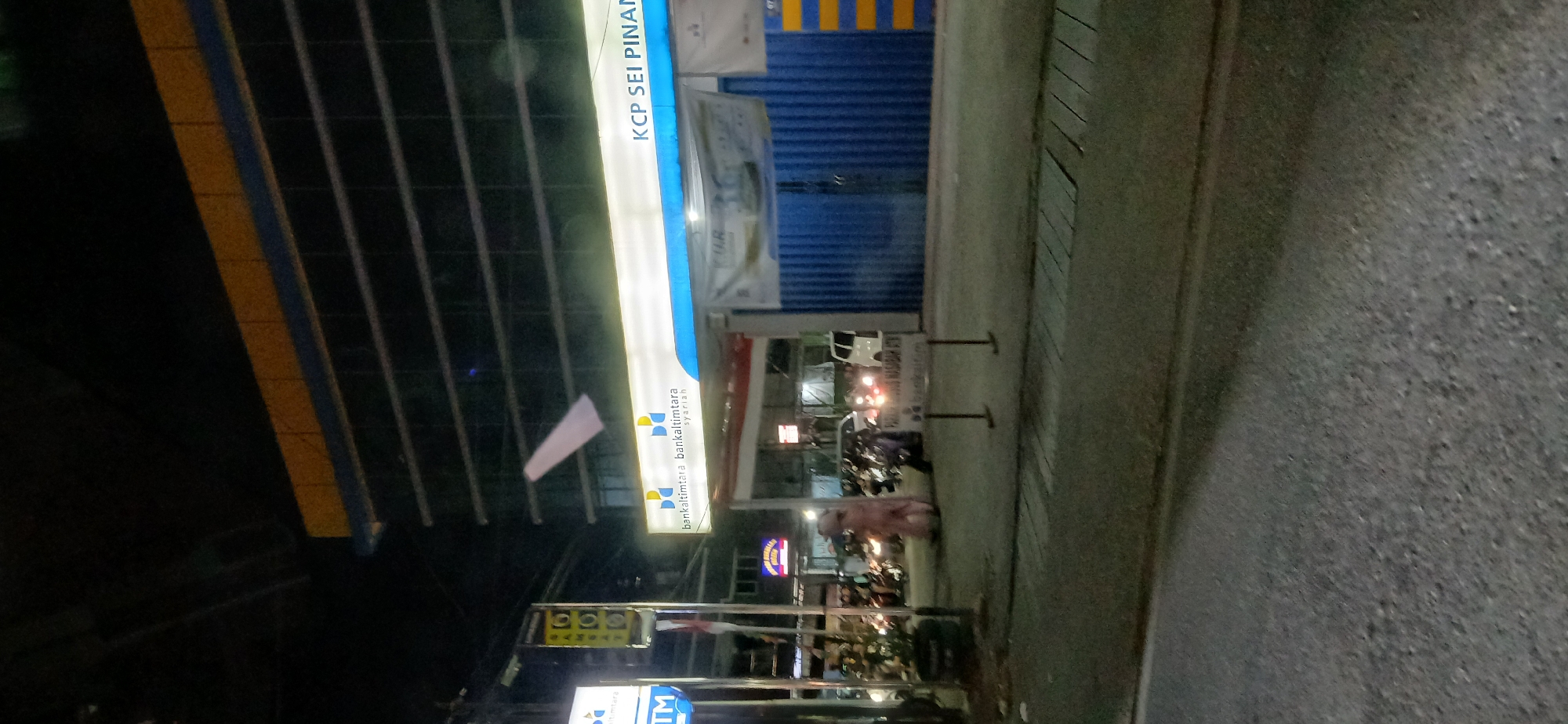 The image depicts the exterior of a Bankaltimtara Syariah branch at night, specifically located in Sei Pinang, Samarinda, Indonesia. The building features a modern facade with a prominent yellow and blue striped awning extending over the entrance. A large, illuminated sign reads "bankaltimtara bankaltimtara syariah" and "KCP SEI PINANG". Several vehicles, including motorcycles and cars, are parked in front of the bank, and some individuals are visible near the entrance. A white banner hangs above the entrance, partially obscured by a white, kite-like object suspended in the air. The surrounding area appears to be a street, with a road in the foreground and sidewalks. The lighting suggests it is nighttime, with artificial lights from the building and street illuminating the scene.