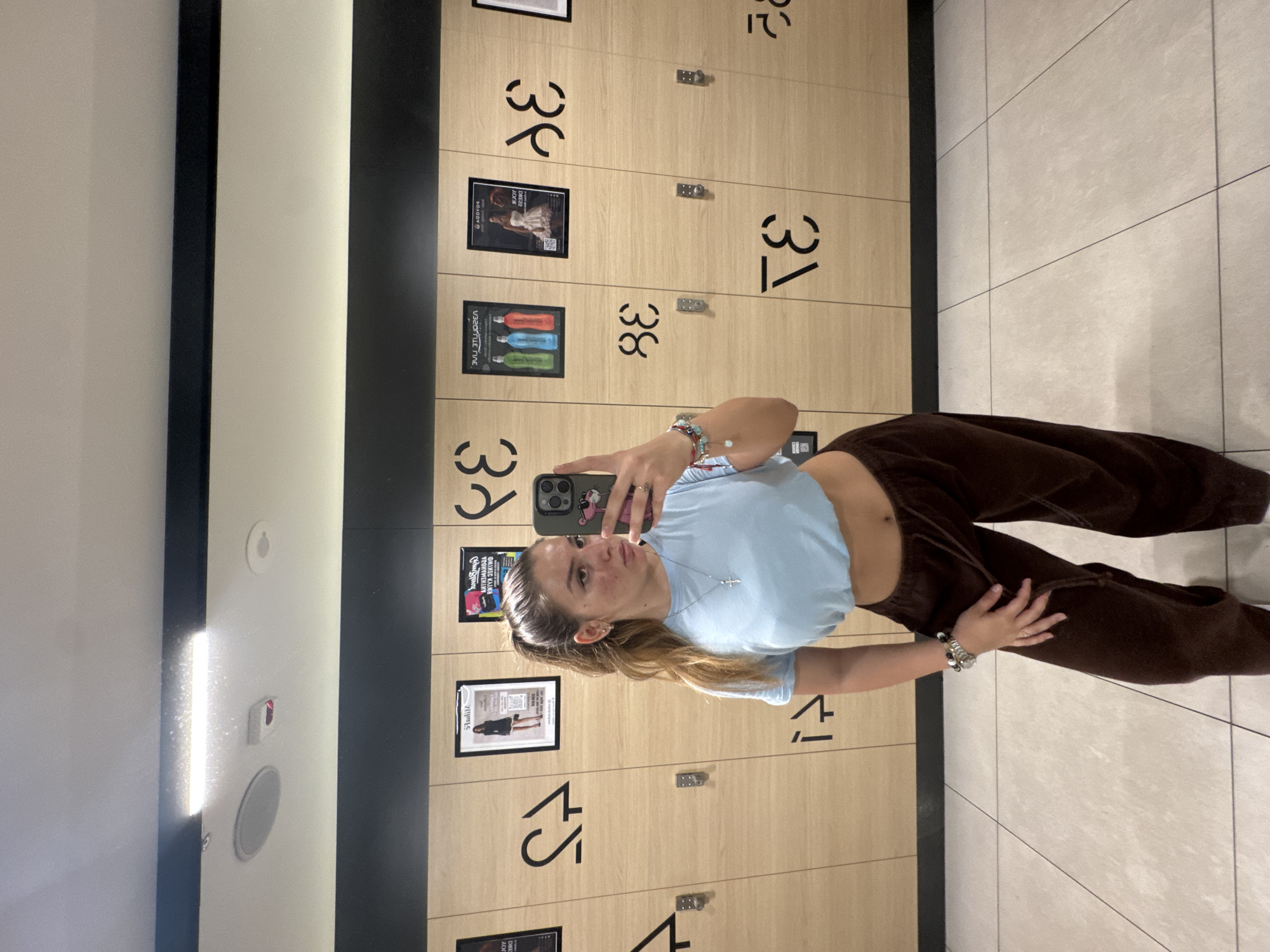 The image depicts a young adult female taking a mirror selfie in what appears to be a modern changing room, likely within a gym or fitness center located in Sofia, Bulgaria.
The subject, a female with light brown hair tied in a high ponytail, stands facing her reflection. She wears a light blue cropped t-shirt, exposing her midriff, and loose-fitting dark brown sweatpants. Her right hand holds a smartphone with a pink panther case, positioned to capture her reflection. A silver-colored cross pendant hangs around her neck, and multiple beaded and metallic bracelets are visible on both wrists. A watch is on her left wrist, and a ring is on her right hand. Her facial expression is neutral, looking directly at the camera. There are visible blemishes on her cheeks and forehead.
The background consists of a wall of light-wood-paneled lockers, each featuring stylized dark numerical cutouts (e.g., "43", "44", "38", "37", "33", "32"). Several framed posters are affixed to the locker doors. Notable posters include one with "СОФИЯ ЛАЙВ" (Sofia Live) and images of colorful water bottles, another with "AGOGO" and "ЙОГА ЗА БРЕМЕННИ" (Yoga for Pregnant Women) in Cyrillic, firmly indicating the location is in Sofia, Bulgaria. Other posters include a fashion advertisement with a woman in a black dress and a poster featuring a woman in a white dress. Small metal hooks are present on some locker doors. The lockers are set above a dark, possibly black, horizontal panel. Above the lockers, the wall is light-colored. The ceiling is also light-colored, featuring a long, rectangular LED light fixture, an oval-shaped speaker, and a red-glowing smoke detector or sensor. The floor is composed of light-toned square tiles with dark grout lines.