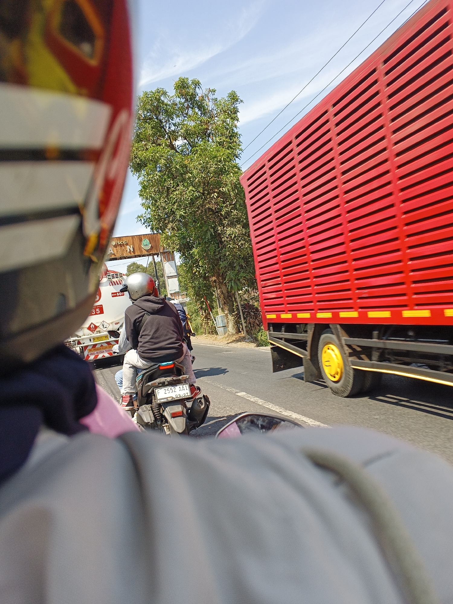 The image depicts a street scene in Sukabumi, Indonesia, likely on a sunny day with a clear blue sky. In the foreground, the perspective is from the back of a motorcycle, with a rider wearing a black hoodie and a silver helmet. The motorcycle is traveling on a paved road, with another motorcycle in front of it, also carrying two people.
To the right of the road, a large, bright red truck with horizontal slatted sides is moving at speed, its yellow wheel prominently visible. The truck appears to be a cargo truck. Further down the road, there's a white truck with visible markings, including the text "ISCG" and "JISCG", suggesting it might be a fuel or chemical tanker. Above the road, a green archway-like structure with signage is visible, also bearing the "ISCG" logo.
The environment features lush green trees along the side of the road, indicating a somewhat rural or suburban setting within the city. The overall impression is a snapshot of daily traffic and transportation in Sukabumi.