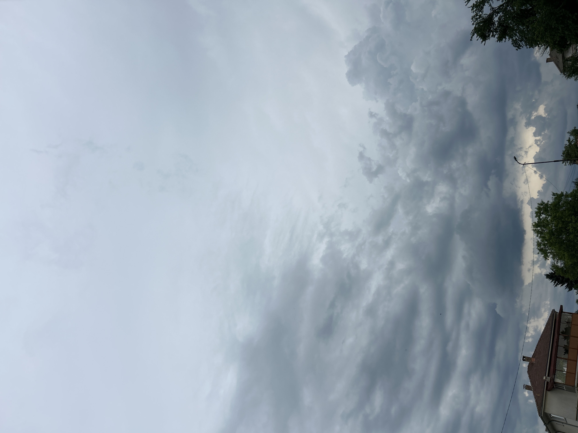 The image shows a cloudy sky over a residential area in Unknown, Unknown. In the foreground, a two-story building with a tiled roof and a balcony is visible on the left. Trees and a streetlamp are also present in the scene. The sky is filled with dark, heavy clouds, suggesting an overcast or stormy day.