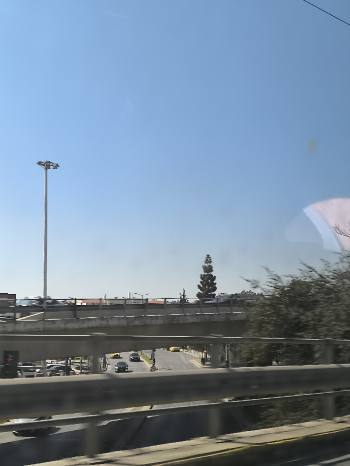 The image shows a highway overpass in the city of Nikea, Greece, taken at latitude 37.977, longitude 23.668. The view is from a vehicle travelling on the highway, looking up at the overpass. The overpass is made of concrete and has a metal railing. There are several vehicles driving on the highway below, including a yellow car, a black car, and a white car. There are also some trees and buildings in the background. The sky is clear and blue. The road surface has a yellow line on the edge. The photo is taken from the passenger side of the vehicle, as the railing is on the driver's side. The view is obstructed by the railing and the windshield. The photographer is not visible in the frame.