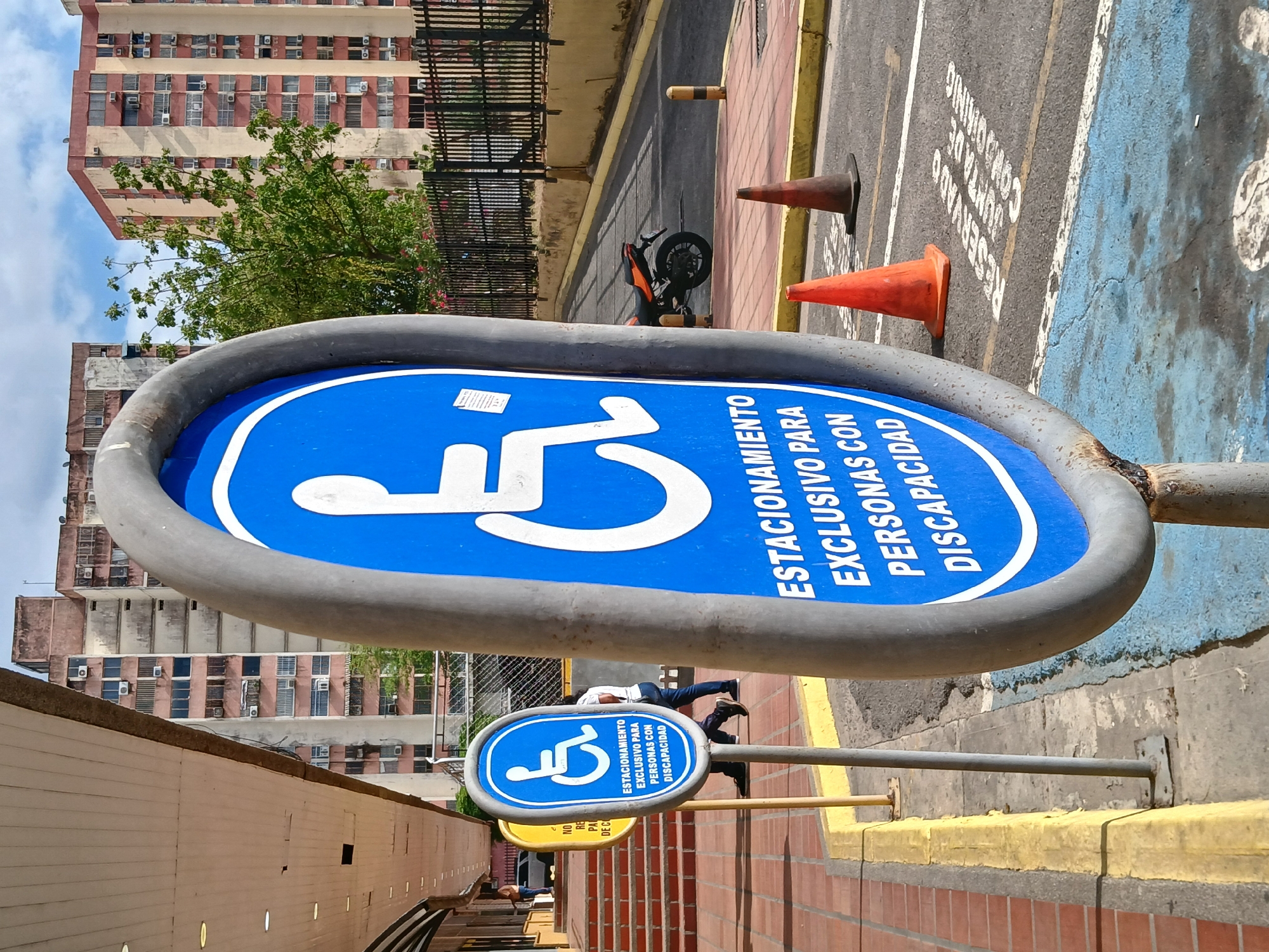 The image displays an outdoor urban environment, likely a street or parking area in Guayana City, Venezuela, under a cloudy daytime sky. The perspective is vertically oriented, capturing a dynamic view of the scene.
The central focus is a large, oval-shaped blue sign indicating accessible parking. It features the International Symbol of Access and the Spanish text "ESTACIONAMIENTO EXCLUSIVO PARA PERSONAS CON DISCAPACIDAD," which translates to "Exclusive parking for people with disabilities." A smaller, rectangular version of this sign is visible further down the street on a pole, displaying the same text.
The ground is a mix of asphalt, red brick paving, and concrete, with yellow painted curbs. On the asphalt, two orange traffic cones are present, along with faded white road markings that include the words "RESERVADO" and partially legible "JUNTA DE COND..." (likely "JUNTA DE CONDOMINIO"). A black and orange motorcycle is parked in the mid-ground near a black metal fence.
In the background, multi-story residential or commercial buildings with numerous windows and visible air conditioning units rise against the sky. Green tree foliage is visible between the street level and the buildings. Two individuals are present in the scene; one person in a white shirt and blue pants stands near the smaller disability sign, while another is partially visible further away. No specific activity beyond being present in the urban setting is discernible for them. A long, light-colored wall runs along the left edge of the frame.