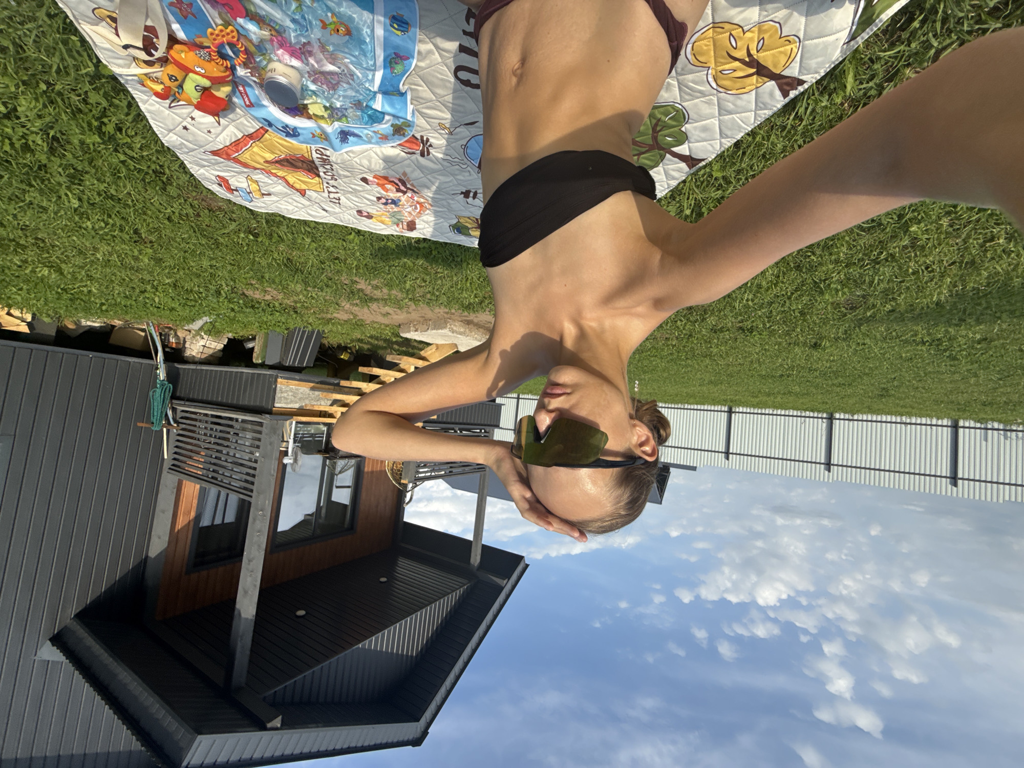 In a sun-drenched outdoor scene in Podshivalovo, Russia, a young woman is captured in a selfie from the chest up. She is seated on a patterned blanket spread across a lush green lawn. She wears a black bandeau bikini top and dark bikini bottoms, and large, mirrored sunglasses that obscure her eyes. Her hair is pulled back in a bun at the nape of her neck. Her left arm is raised, with her hand resting on her forehead. To her right, a modern, dark grey house with wooden accents is visible, featuring a porch with wooden railings. A white metal fence borders the property in the background, with a bright blue sky and scattered white clouds overhead. The blanket beneath her is adorned with colorful graphics, including what appears to be children's toys and a baby bottle.