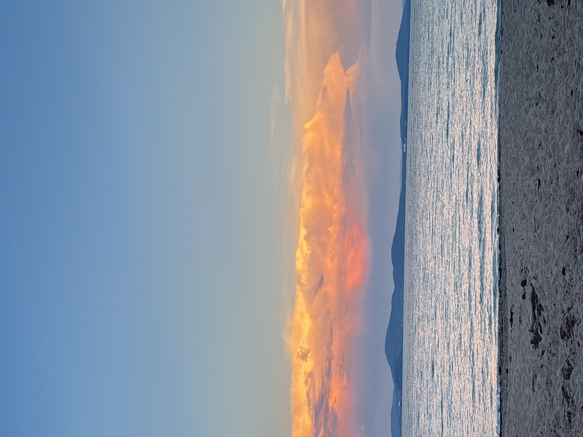 The image depicts a serene seascape at dusk, likely from the coast of Burgas, Bulgaria. The foreground is dominated by a sandy beach, with subtle textures and scattered debris. Beyond the beach, a calm body of water stretches to the horizon, reflecting the soft light of the setting sun. The middle ground features a silhouette of distant, low-lying mountains, their shapes softened by the twilight. Above the mountains, a band of vibrant clouds is illuminated by the last rays of sunlight, displaying hues of orange and pink. The sky above the clouds transitions to a clear, soft blue, indicating the encroaching night. There are no people or animals visible in the scene. The overall atmosphere is peaceful and tranquil, characteristic of a sunset over the sea.
