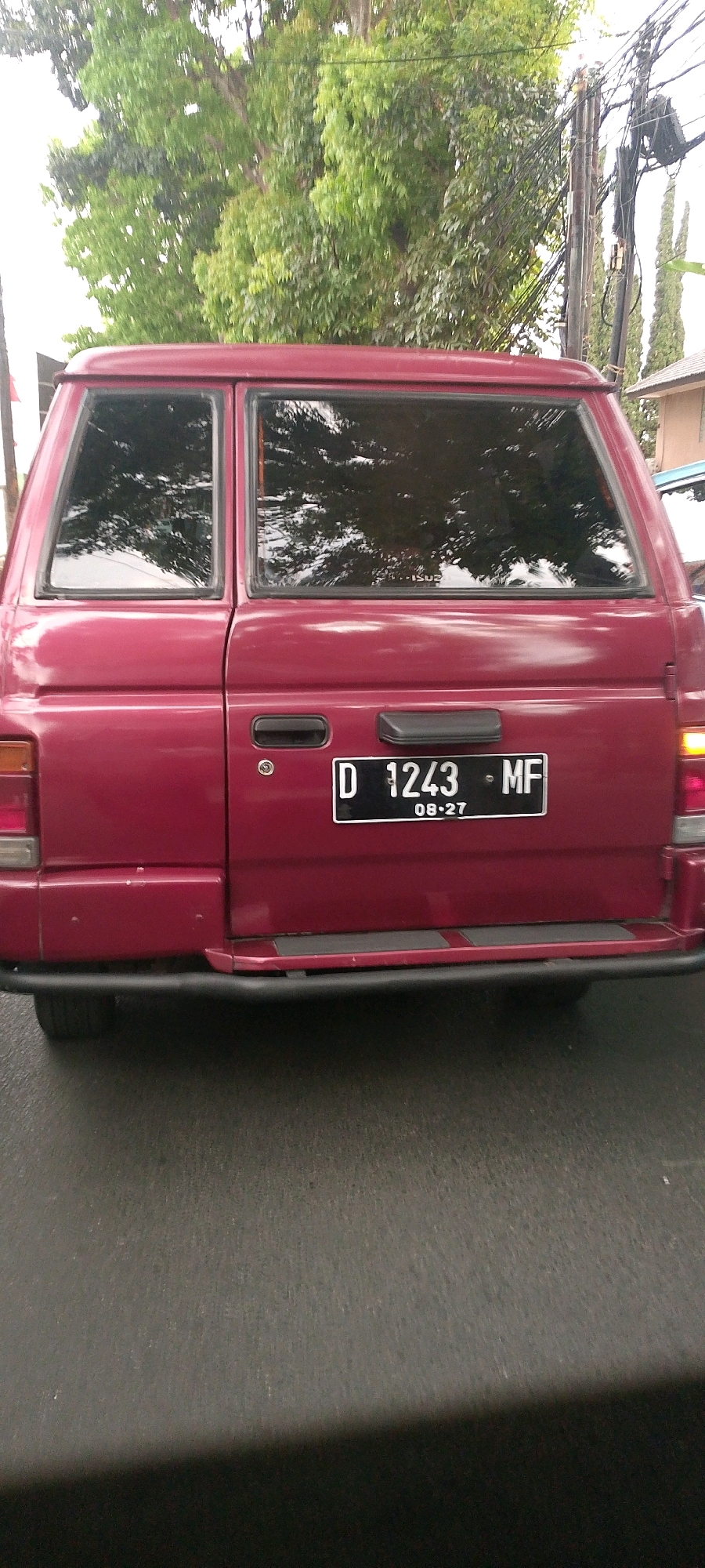 The image shows the rear of a maroon van, likely an Isuzu Panther, parked on a wet asphalt road. The van has tinted windows reflecting trees and the sky. A license plate with white lettering on a black background is visible, reading "D 1243 MF" and "08-27". The background includes trees, utility poles with many wires, and a building to the right. The scene appears to be in an urban environment, possibly in Bandung City, Indonesia, given the license plate prefix "D". There are no people or any visible actions taking place. The lighting suggests it is daytime.