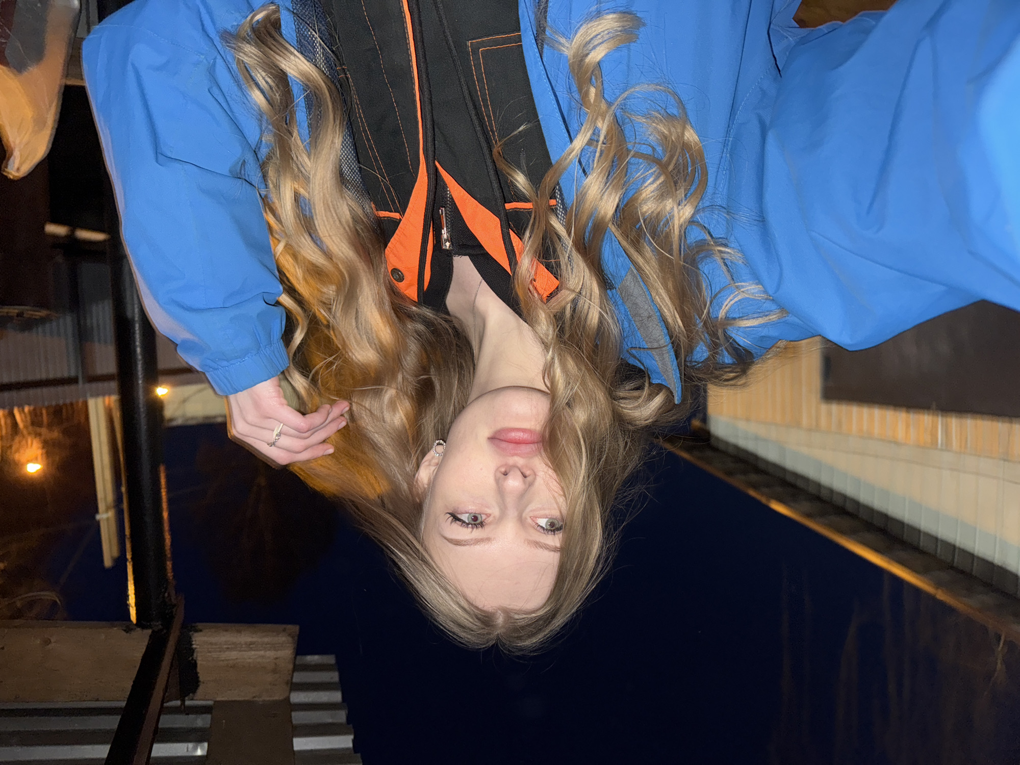 A female with blonde hair is depicted in an upside-down photograph. She is wearing a blue jacket over a black jacket with orange accents. Her right hand is visible, adorned with rings. The setting appears to be outdoors at night, possibly in Perm, Russia, indicated by the architectural style and city layout visible in the background, showing building facades, stairs, and indistinct lighting. The image is taken from a slightly elevated angle.