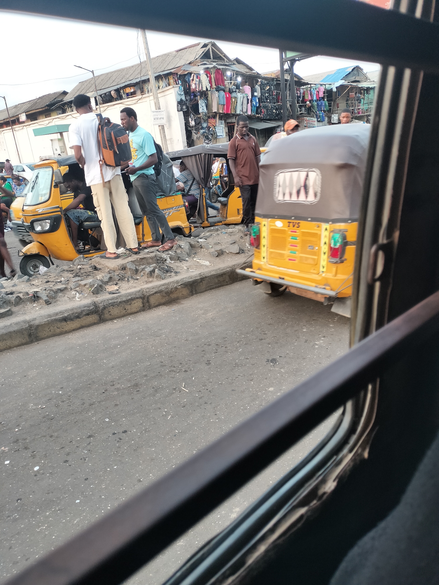 The image shows a busy street scene in what is likely Lagos, Nigeria. Several yellow auto rickshaws (tricycles), a common form of public transportation, are visible, partially obstructed by the view through a window. People are standing near the rickshaws, possibly waiting or negotiating fares. A row of shops selling clothing is visible in the background. The road is paved and appears to be in relatively poor condition. The overall impression is that of a bustling commercial area.