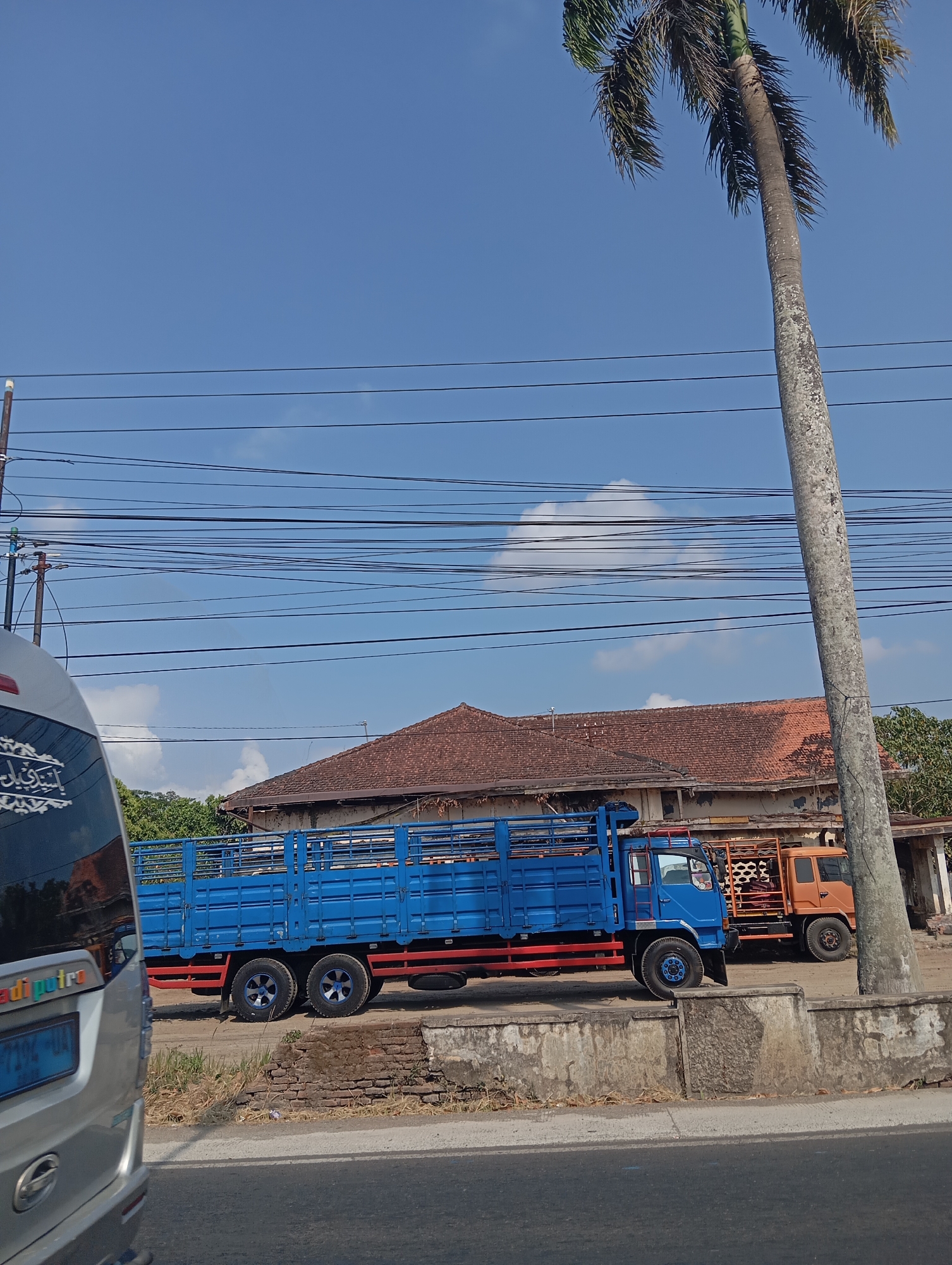 The image captures a street scene in Sokaraja, Indonesia, under a clear blue sky with scattered clouds. A tall palm tree dominates the right side of the frame. In the foreground, the rear of a silver vehicle, possibly a van or bus, is visible on the left, with some text and decorative elements on its back. Further back, a bright blue flatbed truck with a wooden railing is parked, its tires resting on a low concrete wall. To the right of the blue truck, a smaller orange truck is also parked. Behind the trucks, a building with a terracotta-tiled roof and a weathered facade is visible. A network of power lines crisscrosses the sky above the scene. The overall impression is one of a typical roadside environment in a tropical urban setting.
