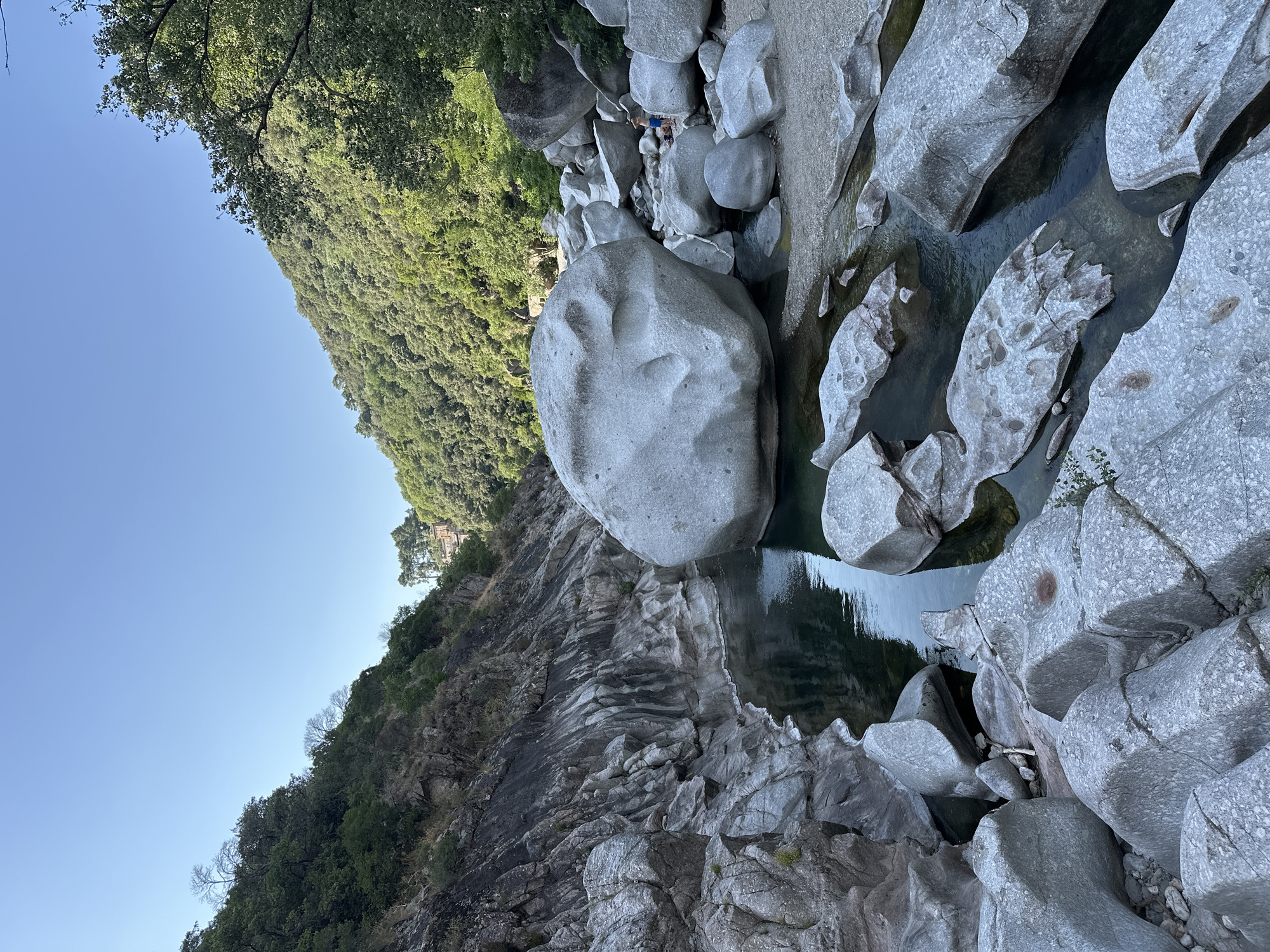 The image shows a wide view of a rocky riverbed with a person visible in the distance. The river is shallow and clear, with smooth, rounded rocks of various sizes scattered throughout. The banks of the river are steep and covered in lush green vegetation. A large, weathered boulder sits prominently in the foreground, its surface etched with natural patterns. The sky is a clear, vibrant blue. A person, wearing blue shorts, is standing on the riverbank near a cluster of rocks. The overall scene depicts a natural landscape, possibly a recreational area.