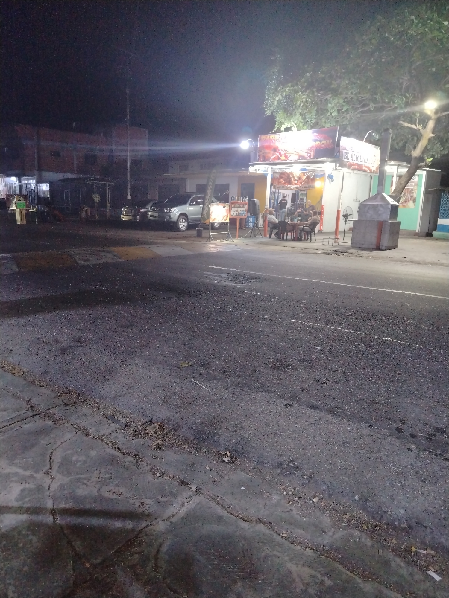 The image depicts an outdoor scene at night in Tinaquillo, Venezuela. A small, brightly lit food stand labeled "El Almendro" is visible, with people seated around a table. A tree is positioned behind the food stand. A street runs in front of the stand. The foreground shows the cracked pavement of the street, while in the background there are several vehicles parked. The buildings in the distance are dimly lit.