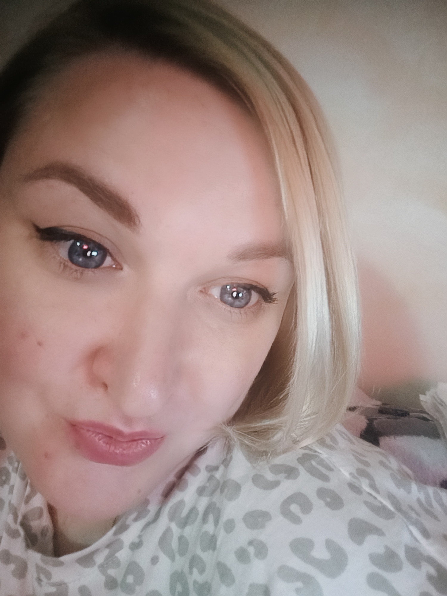 A close-up photograph shows a woman with light skin, blonde hair, and blue eyes. She is wearing eyeliner. Her lips are visible, with a lipstick. She is wearing a white and grey leopard-print top. The location is unknown.