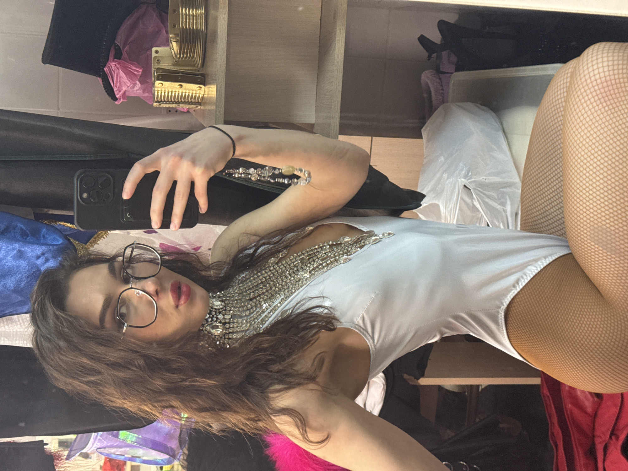 A person with long, wavy brown hair and wearing glasses is lying down and taking a selfie. They are wearing a white bodysuit with an ornate silver and rhinestone necklace or embellishment. The person is also wearing tan-colored fishnet stockings.
The background appears to be a cluttered dressing room or closet space. There are various items of clothing, accessories, and storage containers visible.
There is no information provided about the location being in the city of Kazan, Russia, and the visual information does not suggest this location.