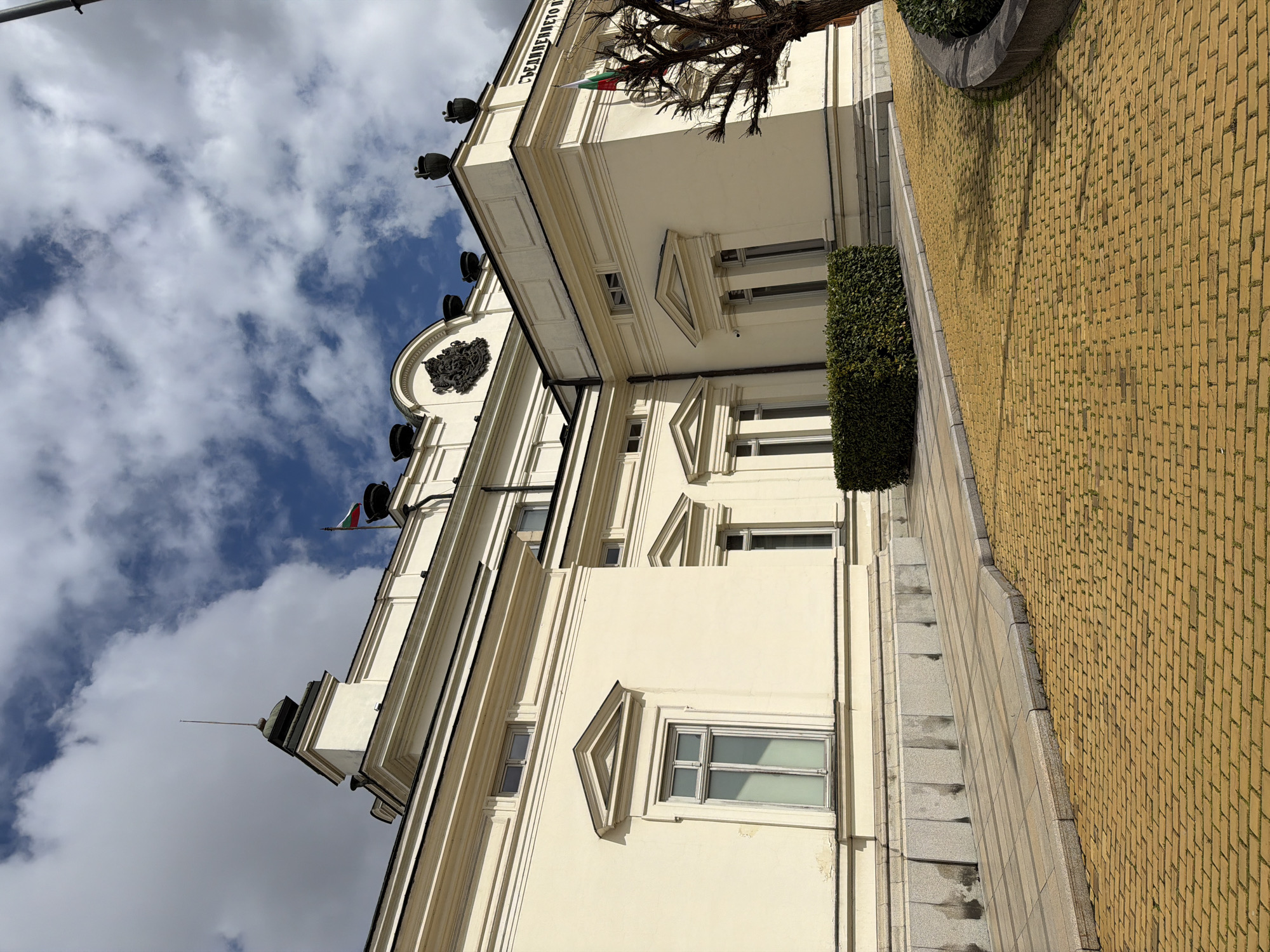 The image captures the National Assembly of Bulgaria in Sofia, Bulgaria. The building is a classical structure with a light-colored facade and detailed architectural elements such as triangular window pediments and a prominent cornice. A Bulgarian flag is visible on the roof. The sky is partially covered with clouds. The sidewalk is paved with yellow bricks. A small bush is on the sidewalk. A bare tree is visible.