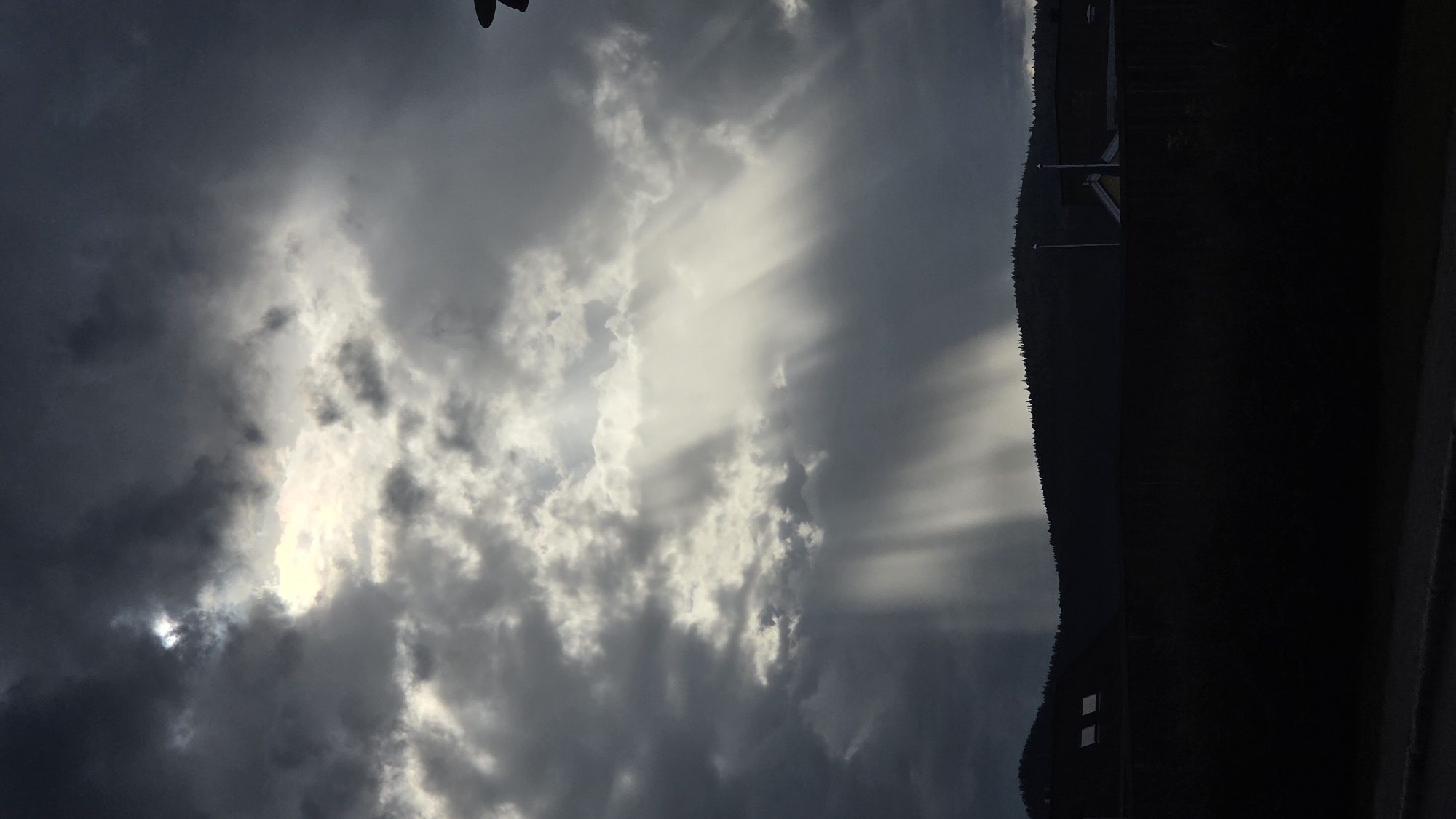 The image depicts a dramatic sky with light rays piercing through dark, heavy clouds over a silhouetted hillside. The sun's rays, known as crepuscular rays, fan out downwards from a bright area in the clouds, illuminating parts of the sky and casting shadows. The hillside is covered with trees, and the faint outline of buildings and structures can be seen along its base. The foreground is largely dark, with a hint of a road or pavement. The scene conveys a sense of impending or passing weather, with a strong contrast between light and shadow. The overall impression is one of natural beauty and atmospheric intensity. Based on the lighting conditions and cloud formations, this scene is likely captured in the city of Tynset, Norway, during a time of day where sunlight interacts with atmospheric moisture and cloud cover to create a striking visual effect.