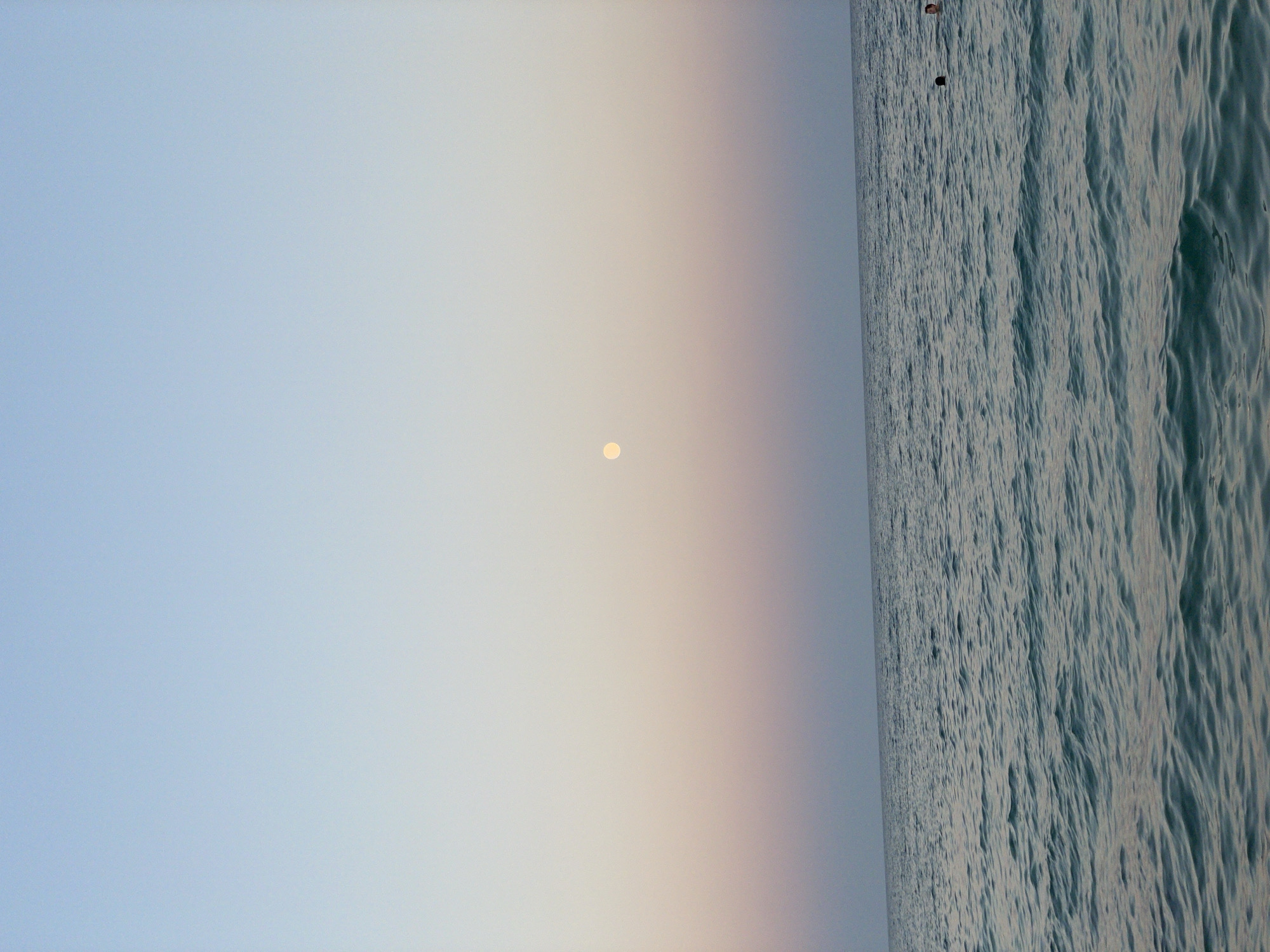 The image depicts a serene seascape at dusk, likely near Trikomo, Northern Cyprus. A pale, full moon is visible in the upper portion of the frame, situated against a gradient sky that transitions from a pale blue at the top to soft hues of pink and peach near the horizon. The sea occupies the lower half of the image, with gentle waves creating a textured surface. Two individuals are visible in the water, one with their head above the surface and the other seemingly further out, their presence suggesting recreational activity. The overall atmosphere is tranquil and peaceful.