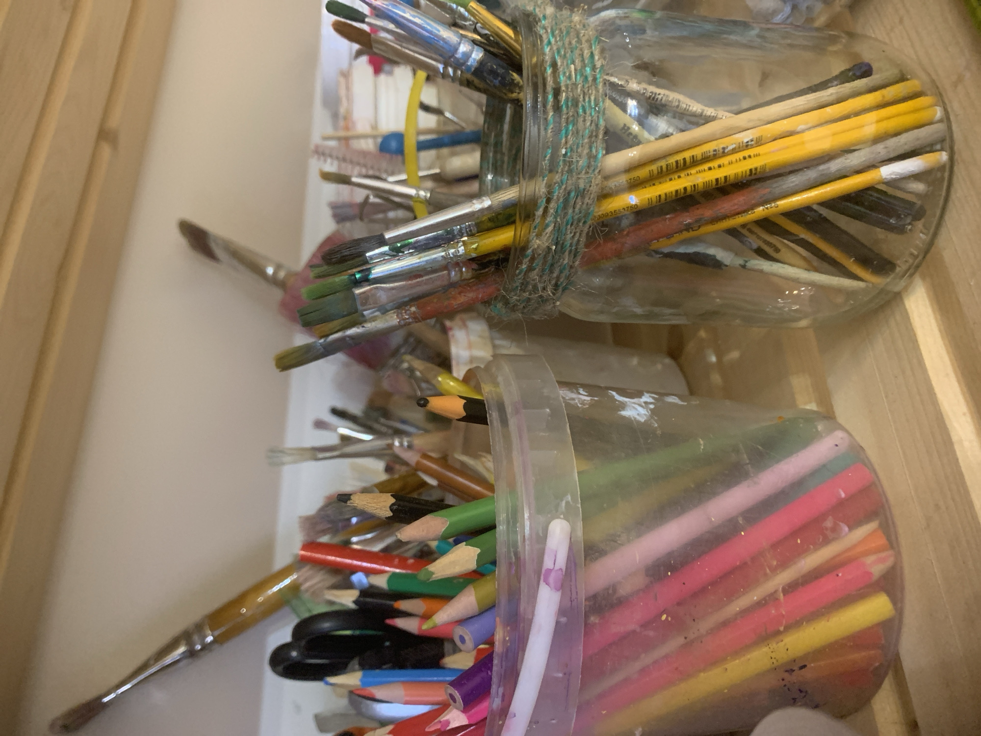 The image shows two containers filled with art supplies. The top container is a clear glass jar secured with a twine tie, filled with an assortment of paintbrushes. The lower container appears to be a translucent plastic tub, containing colored pencils, paintbrushes, and scissors. The scene is indoors, likely a studio or craft room, given the nature of the items. The background is a light beige color, perhaps a wall or cabinet surface. There is no information in the image that would suggest this is located in Izhevsk, Russia.
