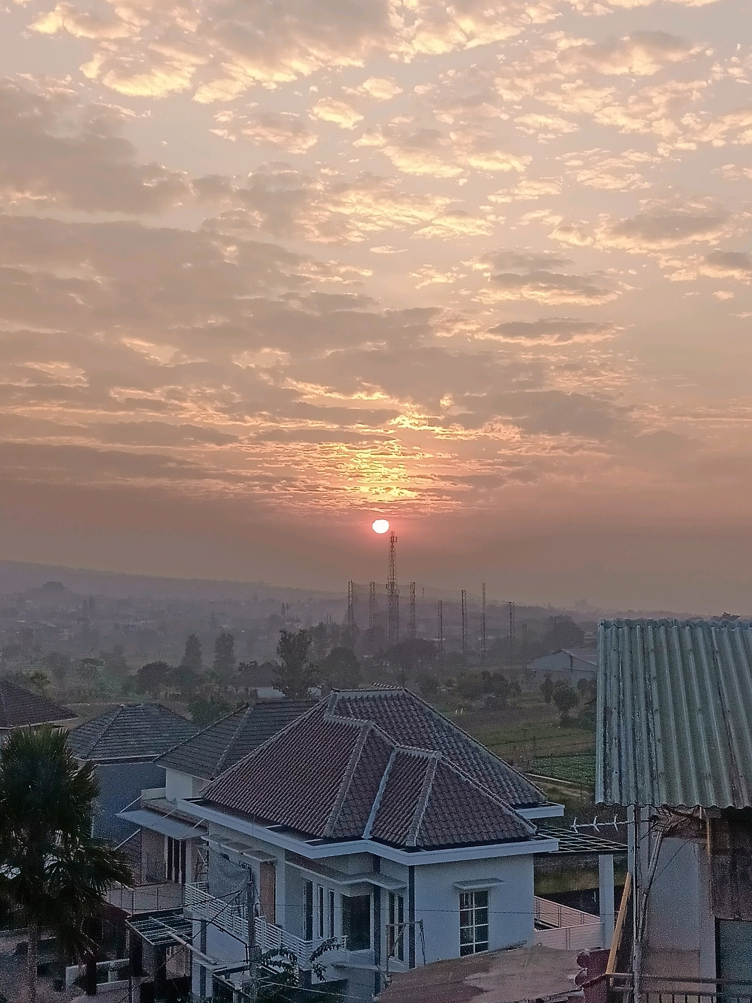 The image captures a hazy sunrise over a residential area in Batu, Indonesia. The sky is filled with soft, orange and pink clouds, with the sun appearing as a bright, small orb peeking through them. In the foreground, several houses with red-tiled roofs are visible. Beyond the houses, a distant landscape includes greenery, a few trees, and several tall communication towers. The overall impression is one of a peaceful, early morning scene in a semi-urban or suburban environment.