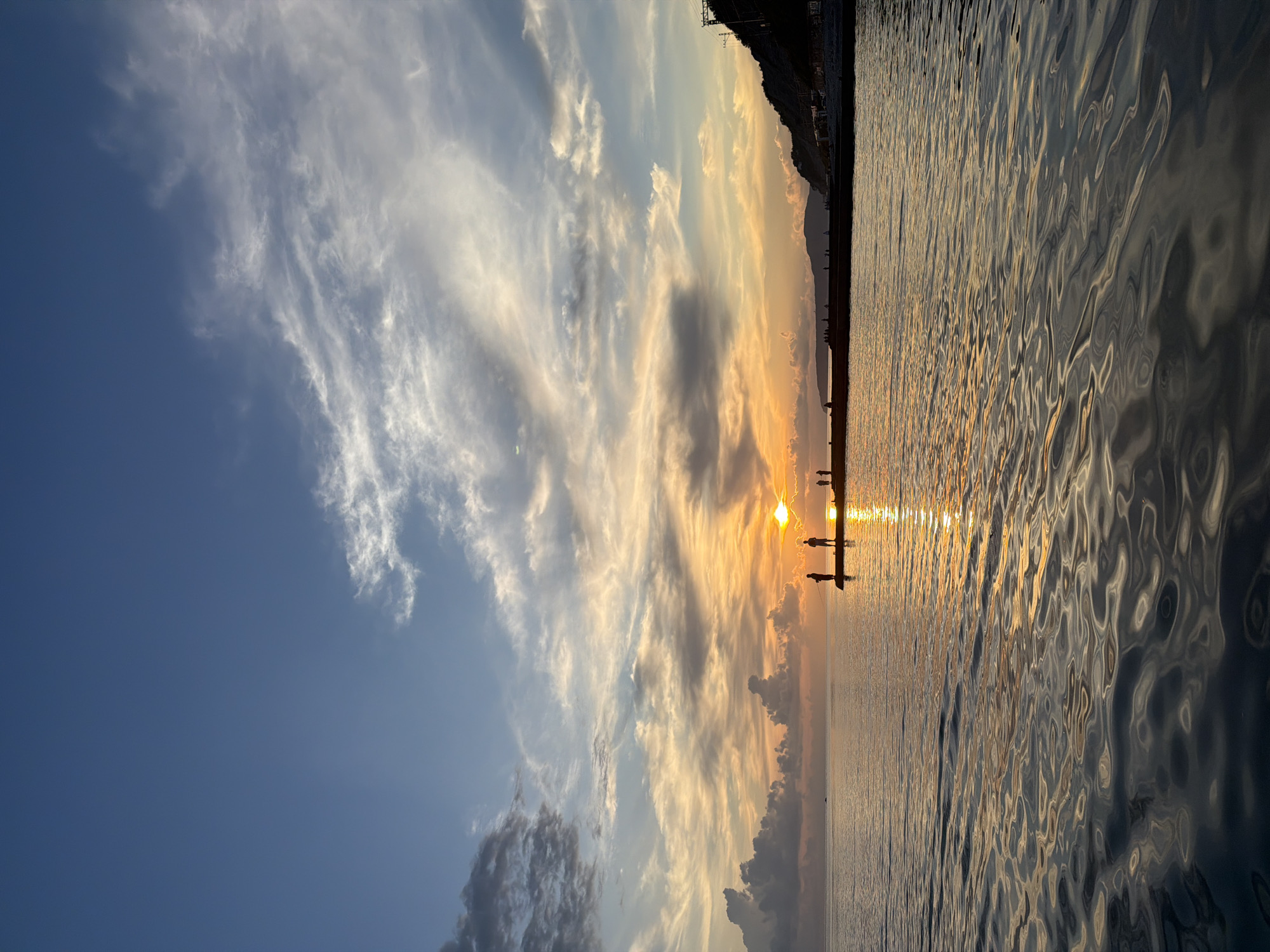 The image depicts a sunset over the Black Sea in Dederkoy, Russia. Silhouettes of several people are visible on a pier that extends into the water. The sun is low on the horizon, casting a warm glow and creating a shimmering reflection on the calm sea. The sky is filled with a mix of blue, orange, and grey clouds. A distant hillside with some structures is visible along the coastline. The water in the foreground shows ripples, suggesting a gentle breeze.