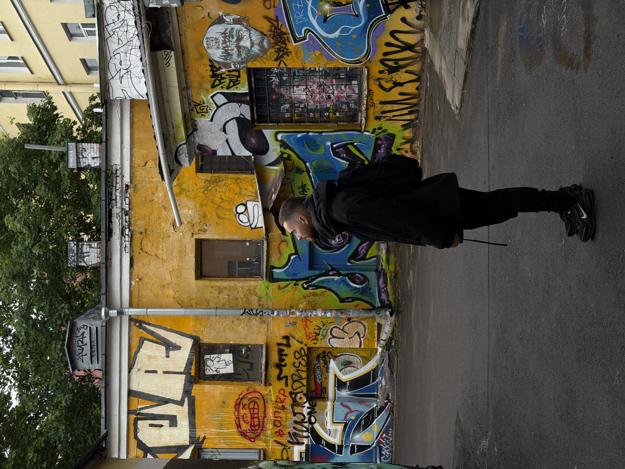 A man stands on an asphalt road beside a building with graffiti, in Saint Petersburg, Russia. The man wears a black jacket and pants, with a backpack and black shoes with white swooshes. He stands perpendicular to the frame and faces left. The building behind him is yellow and covered in graffiti. The graffiti includes images, letters, and tags in blue, black, white, and other colors. There are also several windows on the building. A tree with green leaves is visible on the left side of the frame, partially obscuring the building. Another yellow building is seen above and to the left of the graffiti-covered building.
