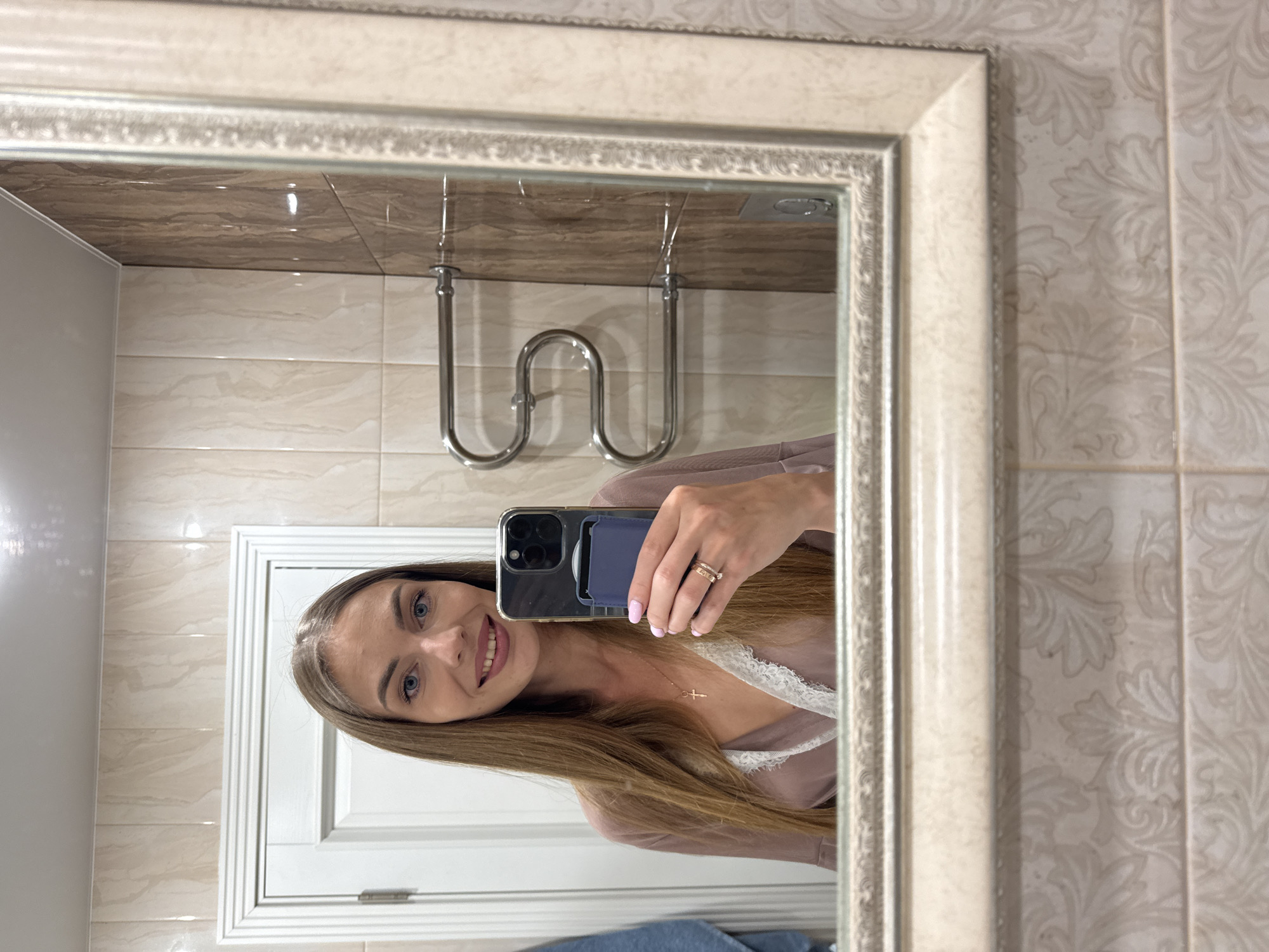 A woman is taking a mirror selfie in a bathroom. She has long, brown hair and is wearing a light brown robe with white lace trim. She is also wearing a necklace with a cross pendant and two rings on her left hand. She is holding her phone in her right hand, and her left hand is resting on her chest. The bathroom has beige and brown tiled walls, a white door, and a chrome towel rack. The mirror has an ornate, light-colored frame. The location is not specified in the image.
