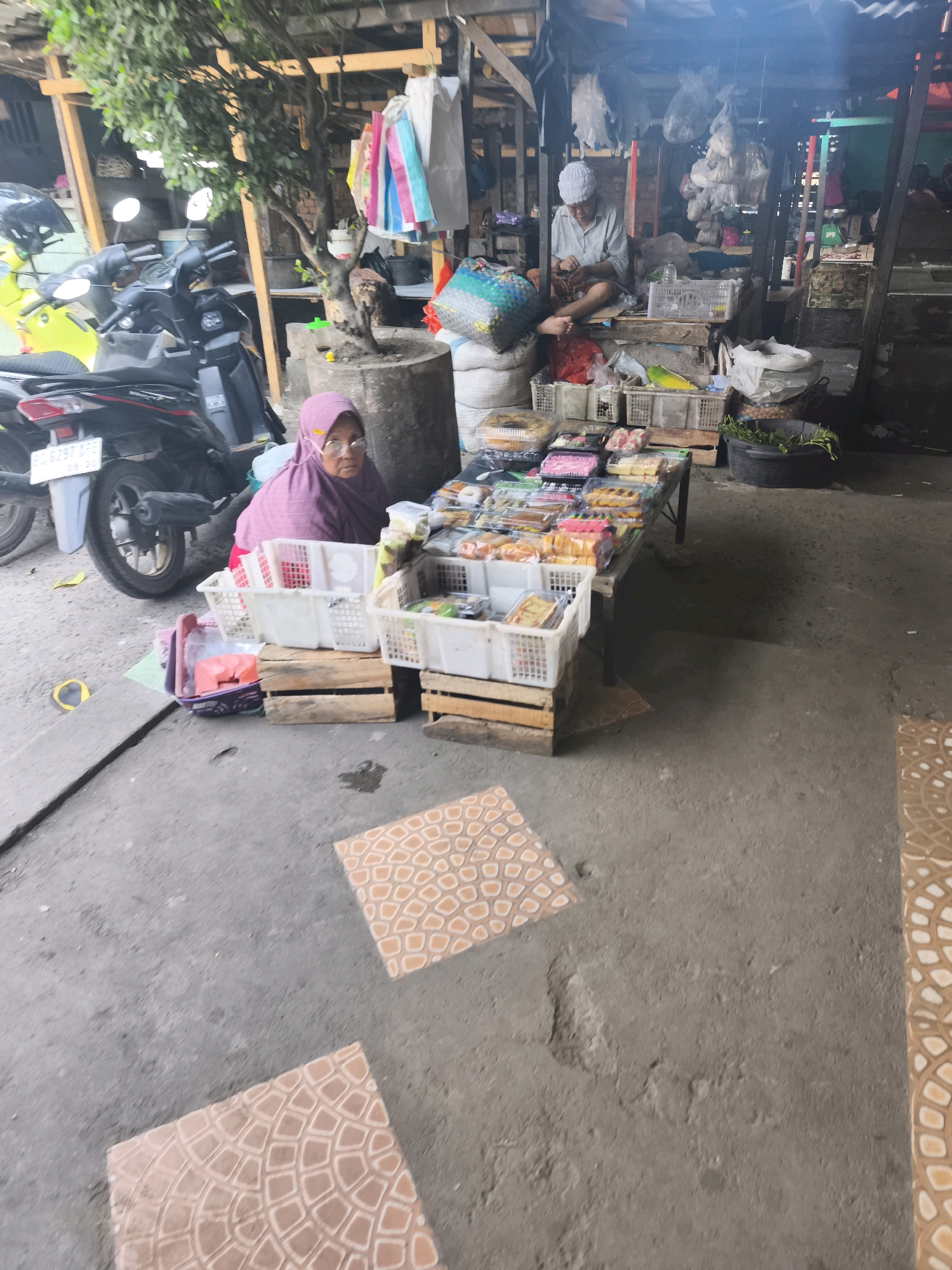The image depicts a street market scene in Palembang, Indonesia, likely in a less developed area based on the rustic structures and unpaved ground. In the foreground, a woman wearing a pink hijab and glasses is seated behind a display of packaged food items. To her right, a man wearing a white cap and a light-colored shirt is engaged in an activity, possibly preparing or arranging goods. The market stall features numerous plastic containers and wooden crates filled with assorted food items, some of which appear to be pastries or snacks. Motorcycles are parked to the left of the scene, suggesting it's a location accessible by vehicle. The environment is characterized by a mix of concrete and dirt ground, with overhead structures and hanging items indicating a makeshift market setup. In the background, more stalls and the general ambiance of a bustling market are visible. The lighting suggests it is daytime, though some areas are shaded.
