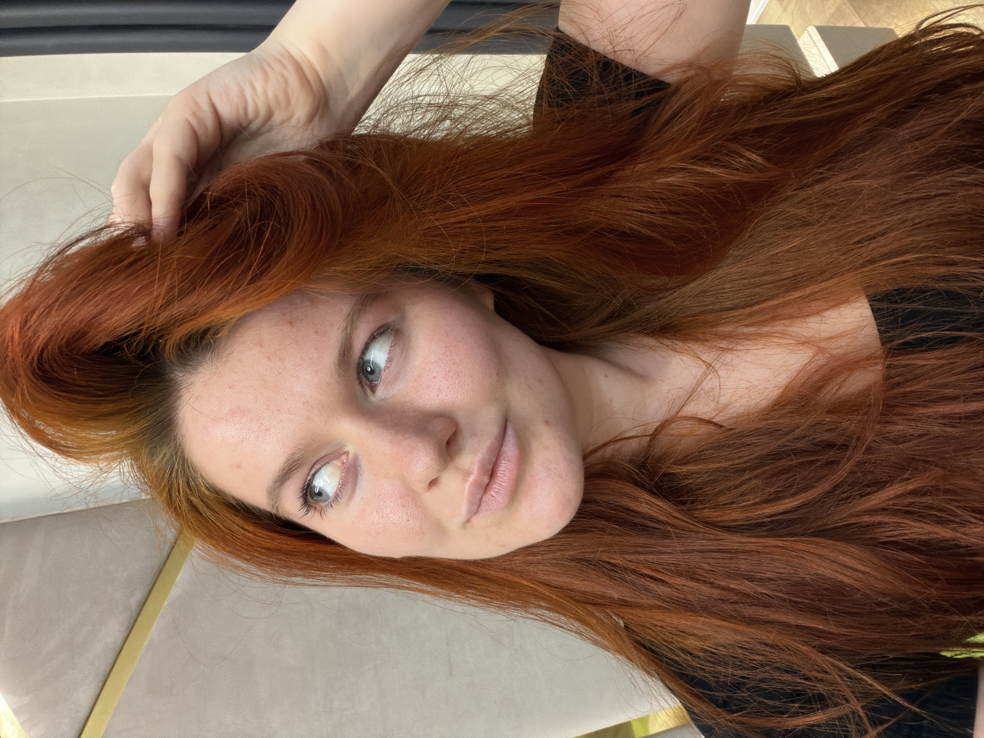 A red-haired woman is looking upwards and to the left, with her right hand touching her hair. She is wearing a black top. The background is a softly lit indoor space with furniture visible. This image was taken in Krasnodar, Russia.