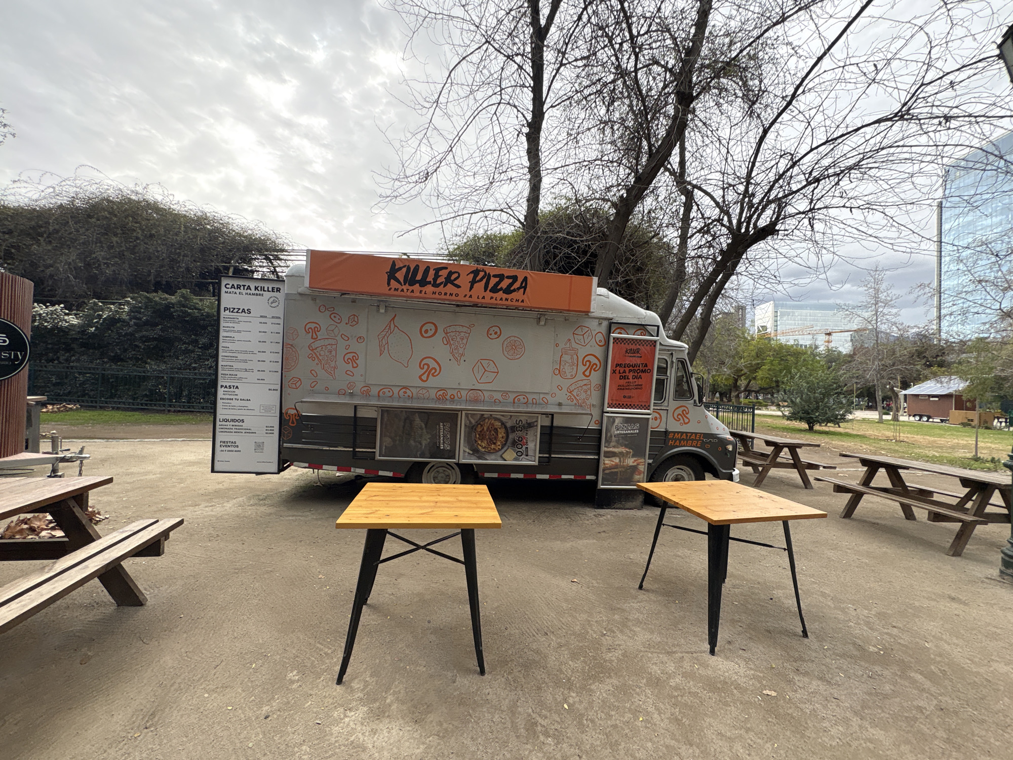 In a park setting in Santiago, Chile, a Killer Pizza food truck is parked. The truck features a large orange banner with the "Killer Pizza" logo and the tagline "#Mata El Horno #A La Plancha". The truck's side is decorated with various cartoonish illustrations of pizza, mushrooms, and lemons. Attached to the side of the truck is a menu board listing "Pizzas," "Pasta," "Líquidos," and "Fiestas/Eventos". Two square wooden tables with black metal legs are set up in front of the food truck, facing the viewer. In the background, several wooden picnic tables and benches are arranged, suggesting a public dining or recreational area. The ground is a sandy, dirt surface. Beyond the park, modern buildings, including a tall glass skyscraper, are visible, indicating an urban environment. The sky is overcast with a mix of clouds. The scene appears to be outdoors during the day.