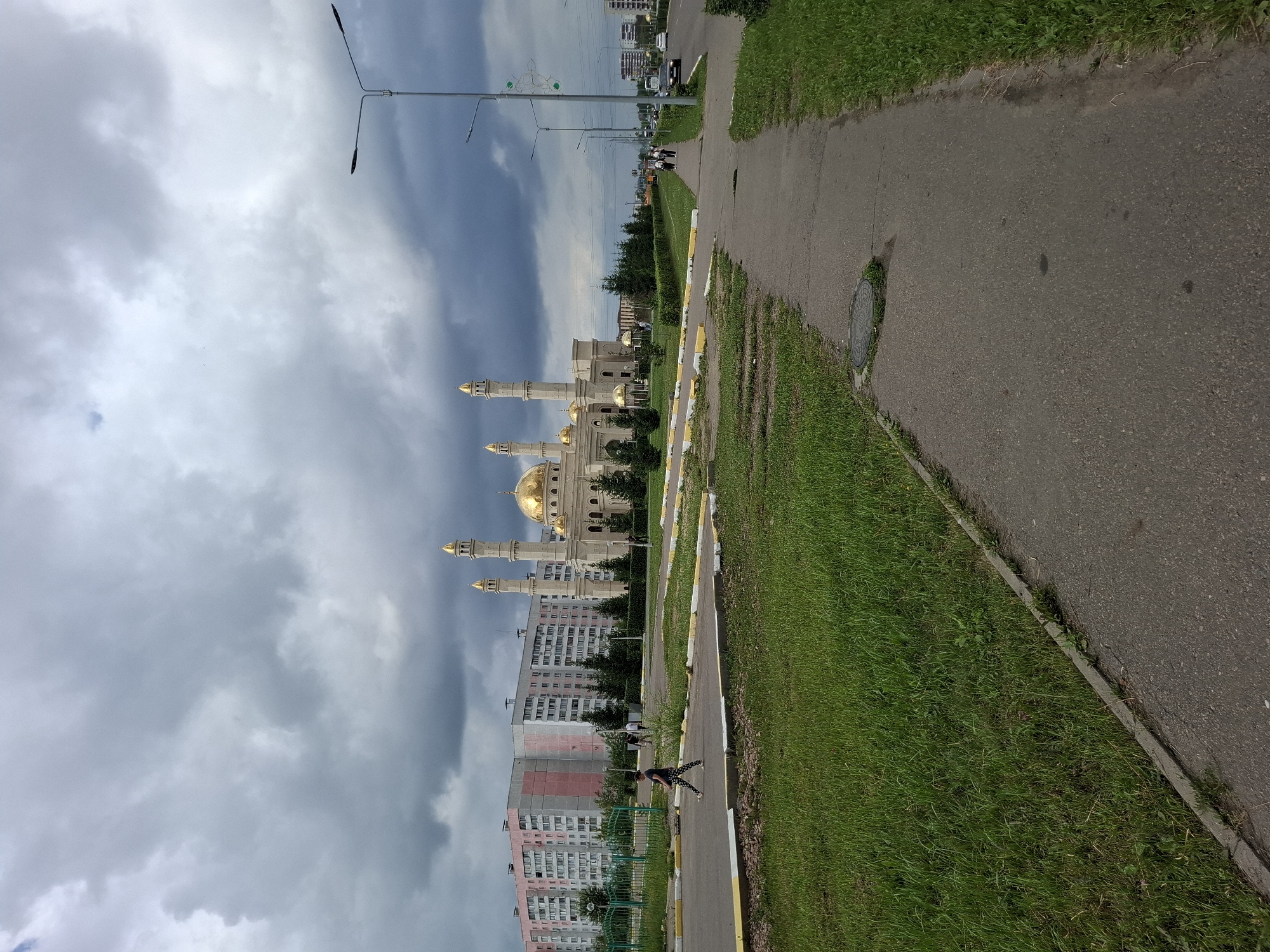 The image shows a view of the Presidential Mosque in Petropavl, Kazakhstan, on a cloudy day. A paved walkway runs diagonally from the lower right to the middle left of the frame, bordered by a strip of grass on the left and right. A woman with dark hair, wearing a dark t-shirt and patterned leggings, walks along the walkway in the foreground, facing left. Behind her, a low green fence separates the walkway from a grassy area. Further in the background, to the right of the fence, stands the Presidential Mosque, a prominent structure with a large golden dome and two tall minarets, also topped with golden spires. Adjacent to the mosque, to its left, are multi-story apartment buildings. Several evergreen trees are planted around the mosque and in front of the apartment buildings. The sky is overcast with dark, heavy clouds. In the distance, on the right side of the image, a street is visible with traffic, including cars and a bus. Lampposts line the street.