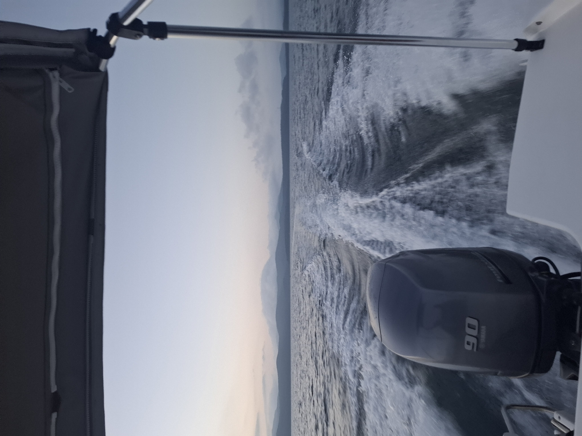 The image depicts a view from the back of a boat moving through the water. In the foreground, a gray outboard motor with "90" and "YAMAHA" visible on it is prominently featured. The boat is creating a white wake behind it as it travels across a body of water. In the distance, a hazy shoreline with hills or mountains is visible under a pale sky with some clouds. The overall impression is of a calm, early morning or late evening on the water. The location is identified as Dunai, Russia.