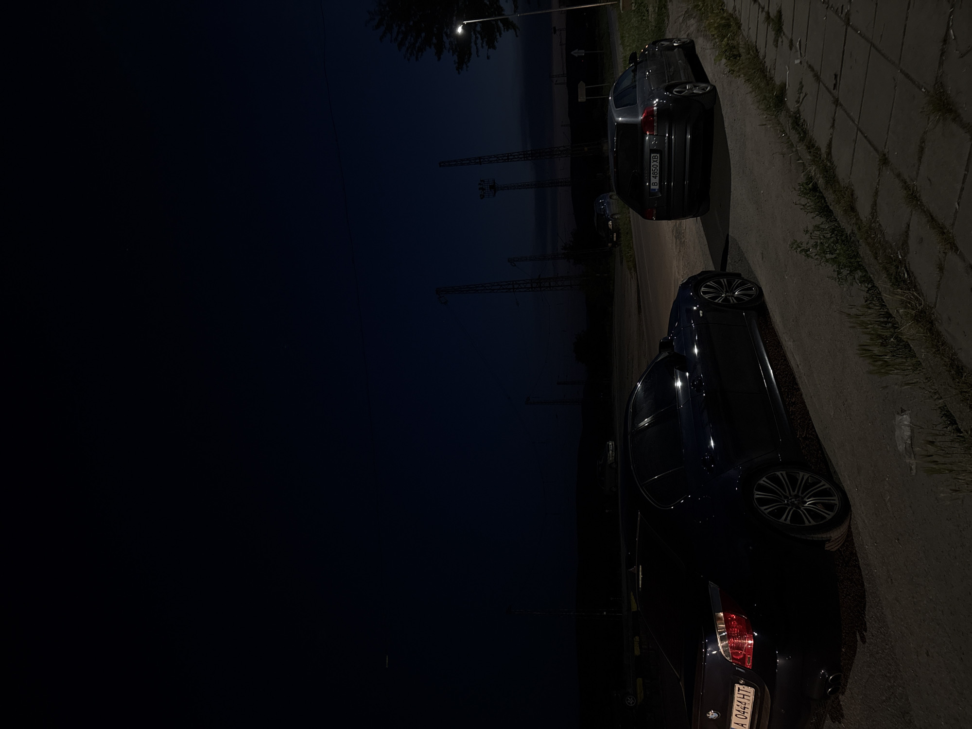 In the city of Dalgopol, Bulgaria, at night, two cars are parked on a dirt road next to a paved area. The car on the left is a dark-colored BMW sedan, seen from its rear quarter, parked at an angle. Its taillights are illuminated. The car on the right is a grey Audi station wagon, parked facing away from the viewer. Its license plate is visible and reads "B 4850 XB". Beyond the cars, industrial-looking towers with lights are visible against a dark sky. A single streetlamp illuminates a portion of the paved area in the foreground. The environment appears to be an outdoor, possibly semi-rural or industrial area, with sparse vegetation growing along the edge of the pavement. The sky is dark, indicating it is either late evening or early morning.
