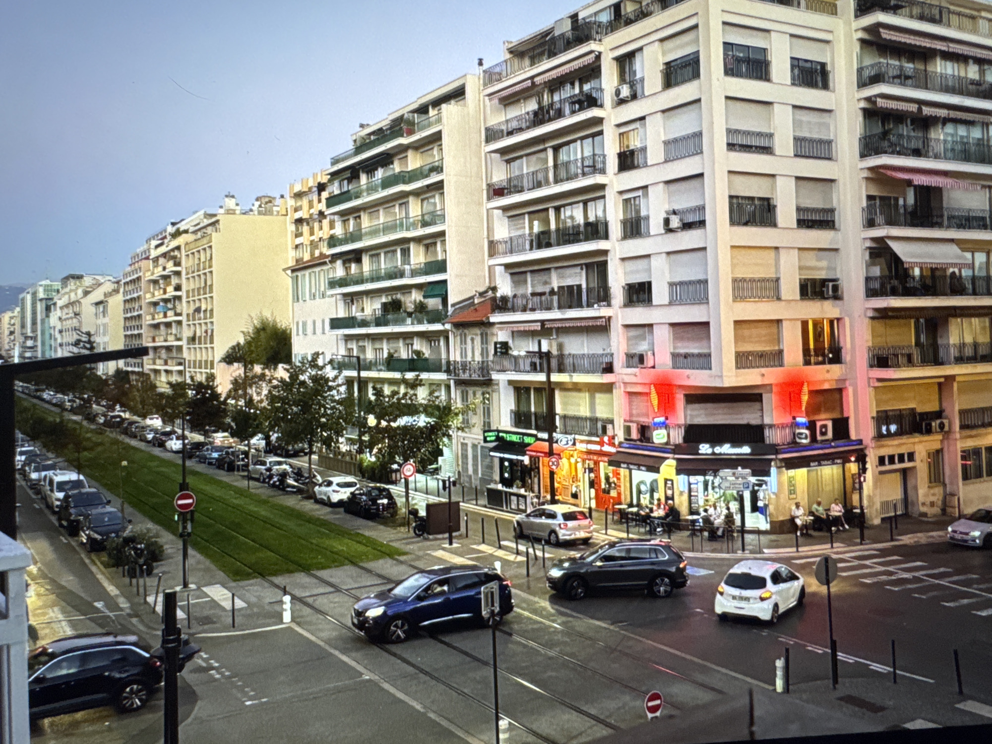 The image depicts a street scene in a European city, likely Nice, France, given the style of architecture and signage, not Porrentruy, Switzerland. In the foreground, a dark blue station wagon drives on tram tracks. To its right, a dark gray SUV is also on the street. Further up the street, a white hatchback turns at an intersection. Across the street, several people are seated at tables outside a cafe labeled "Le Muguet" and "BAR TABAC Presse". The buildings lining the street are multi-story, with balconies and awnings. On the left side of the image, a green median strip separates the tram tracks from a row of parked cars. The sky is overcast.