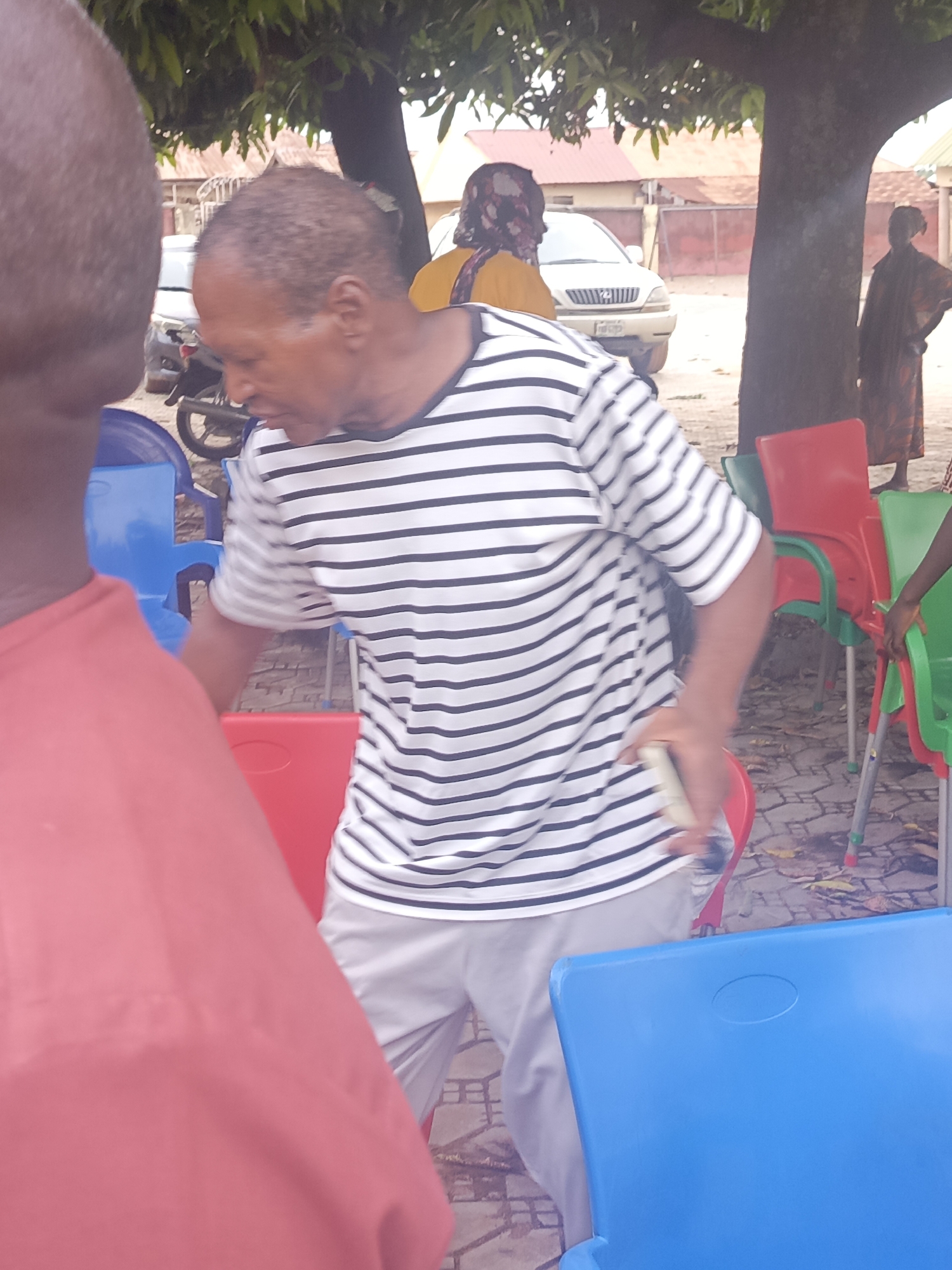 A medium shot of a man in a striped shirt and light gray pants stands among red and blue plastic chairs outdoors. He is looking downward while holding a phone in his right hand. The man is located in Akwanga, Nigeria. Behind him, partially obscured by trees, are a parked white SUV, a parked motorcycle, and other people. One woman is wearing a yellow top and a patterned headscarf, and another is wearing a patterned dress. A person's head and shoulders are visible in the foreground on the left, wearing a red shirt. The ground is paved with a mottled pattern.