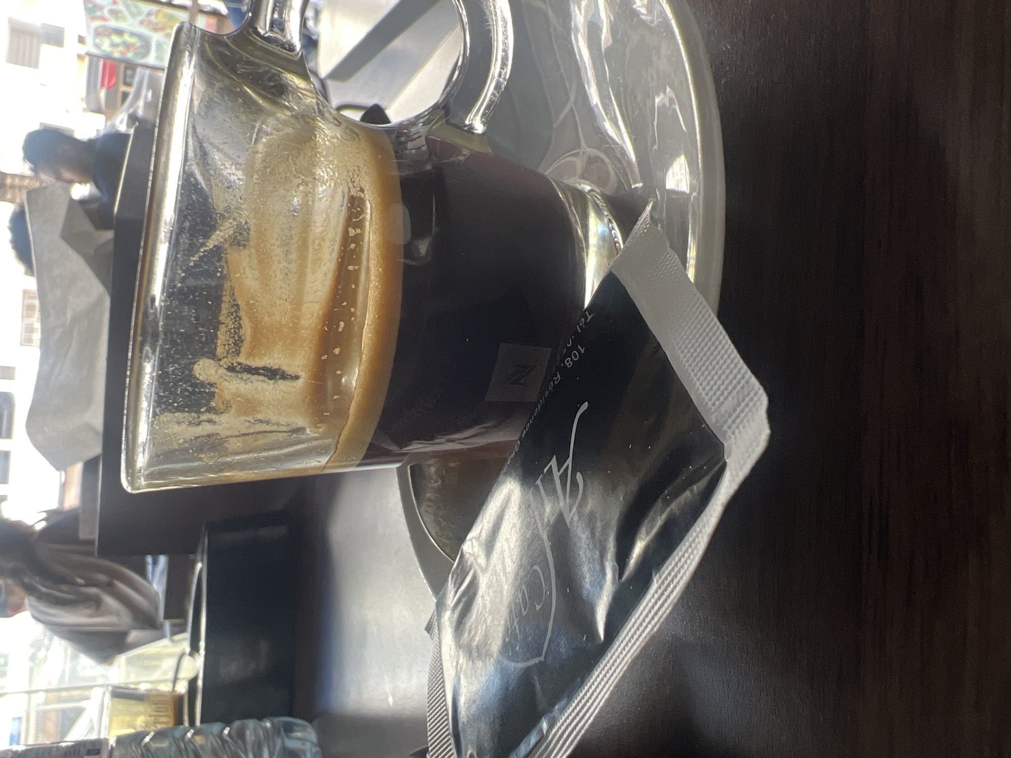 The image captures a close-up, eye-level perspective inside a brightly lit cafe, likely located in Casablanca, Morocco. In the immediate foreground, a dark wooden table surface is visible. Centrally positioned, a transparent glass mug, branded with the "Nespresso" logo, contains approximately half a serving of dark coffee topped with a prominent layer of light brown crema. The mug rests on a white ceramic saucer. To the left of the mug, a sealed black rectangular packet, identified as a sugar or sweetener packet, lies on the table. White text on this packet includes "Café ZA" (stylized logo) and partially visible address details such as "108, R" and "Tel.". A clear plastic water bottle is partially visible on the extreme left. The background is significantly brighter, indicating large windows or an open area. Blurred figures of other individuals are discernible in the background: one person with long, light-colored hair and a light top is partially visible on the left, facing away from the camera; another person with dark hair and a dark top is seen on the right. A crumpled white tissue is also present on a surface in the background. The branding on the sugar packet, "Café ZA," corresponds with known establishments such as "Café Zara" in Casablanca, suggesting the specific urban context. The scene suggests a paused moment during a beverage consumption in a public cafe setting.